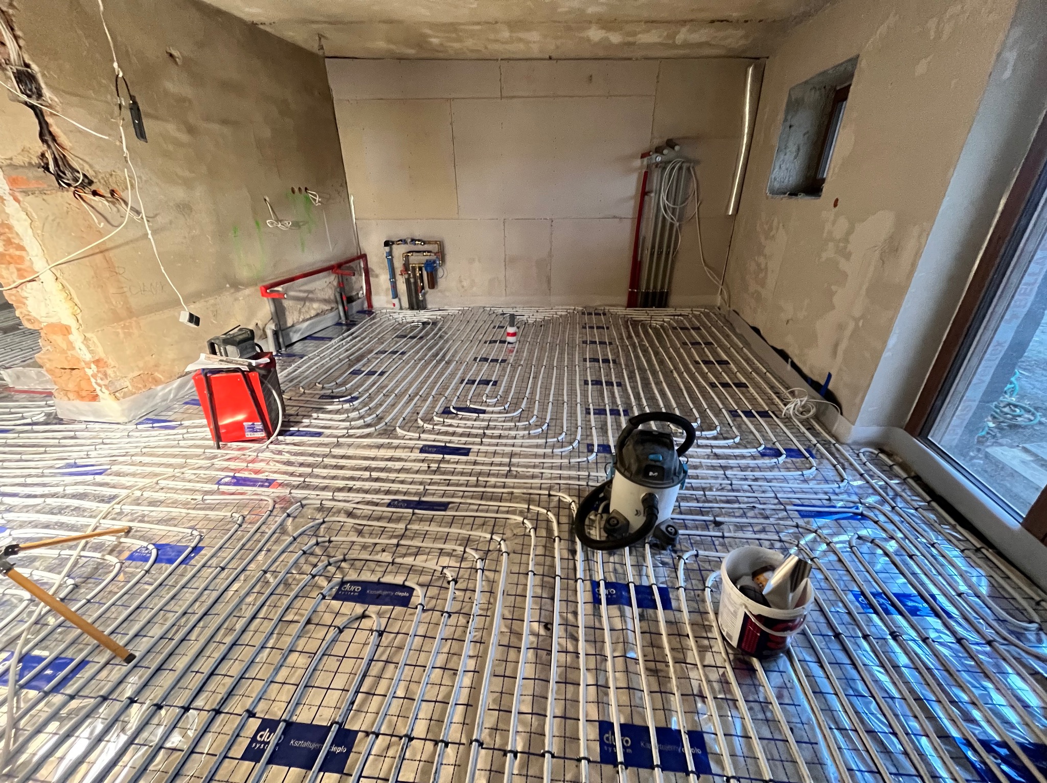 Montaż ogrzewania podłogowego w surowym wnętrzu. Widoczne rury na folii izolacyjnej, narzędzia i przygotowane ściany. Częściowy widok okna.