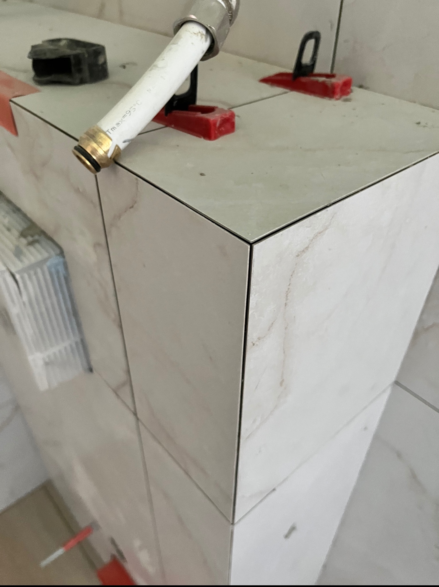 Wykończona obudowa z płytek imitujących marmur, z widocznymi elementami poziomowania i fragmentem rury z metalowym złączem, ukazana z bliska.