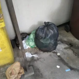 Sterta odpadów w workach i luzem przy ścianie, obok żółty pojemnik. Widoczny fragment brudnej podłogi. Niejednolita kolorystyka i brak estetyki.