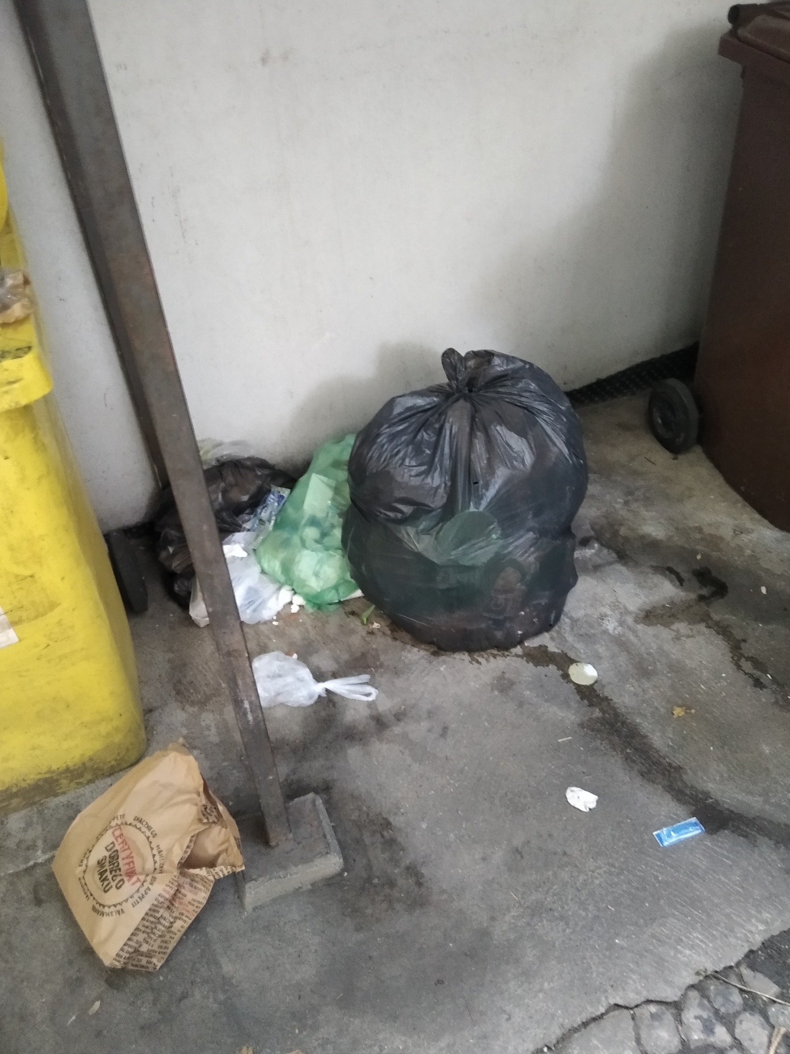 Sterta odpadów w workach i luzem przy ścianie, obok żółty pojemnik. Widoczny fragment brudnej podłogi. Niejednolita kolorystyka i brak estetyki.