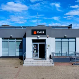 Budynek składu budowlanego Plan B z widocznym logo i otwartymi drzwiami wejściowymi. Na placu zaparkowany samochód dostawczy.