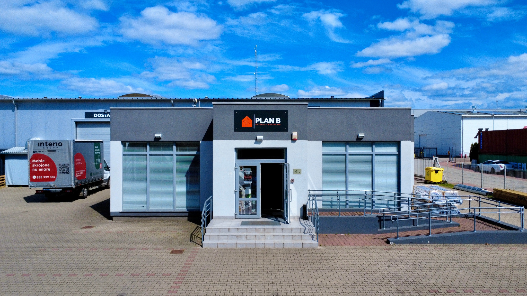 Budynek składu budowlanego Plan B z widocznym logo i otwartymi drzwiami wejściowymi. Na placu zaparkowany samochód dostawczy.