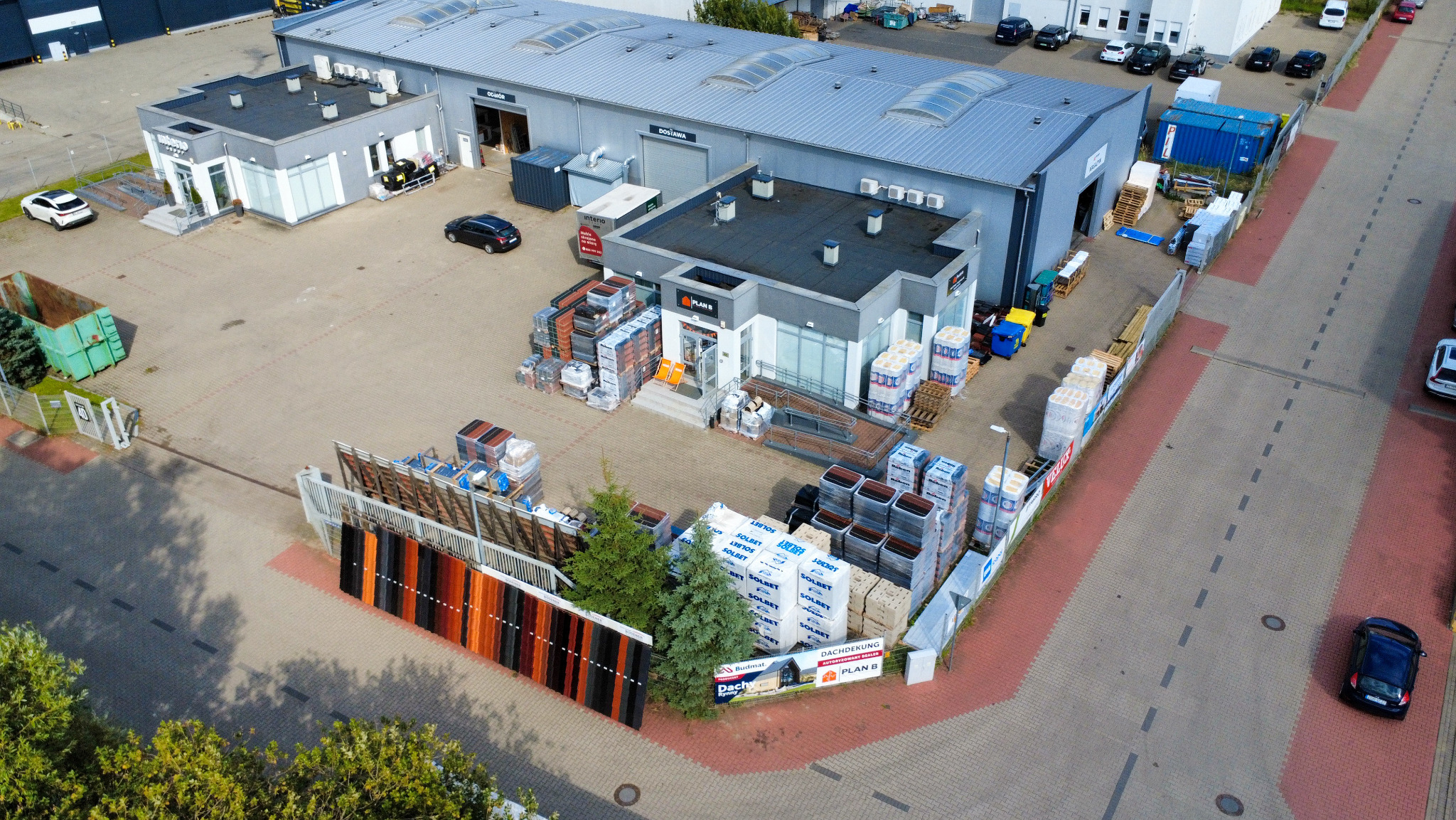 Widok z góry na skład budowlany z ekspozycją dachówek i materiałów izolacyjnych, z budynkami biurowymi i magazynem w tle. Widać też zaparkowane samochody.