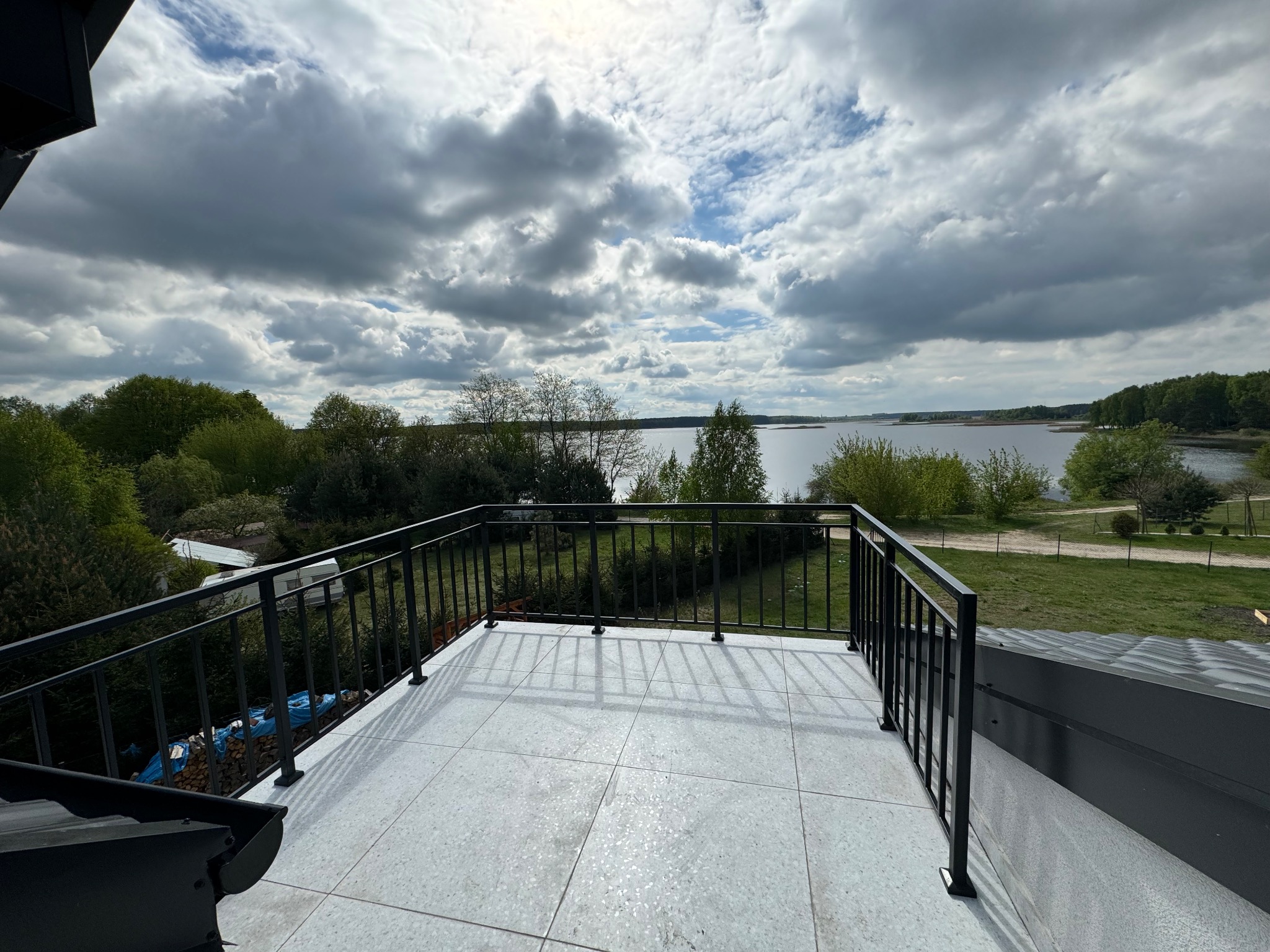 Czarna, metalowa balustrada balkonowa z widokiem na jezioro i zachmurzone niebo. Jasna, kwadratowa płytka podłogowa. Widok z góry, szeroki kadr.