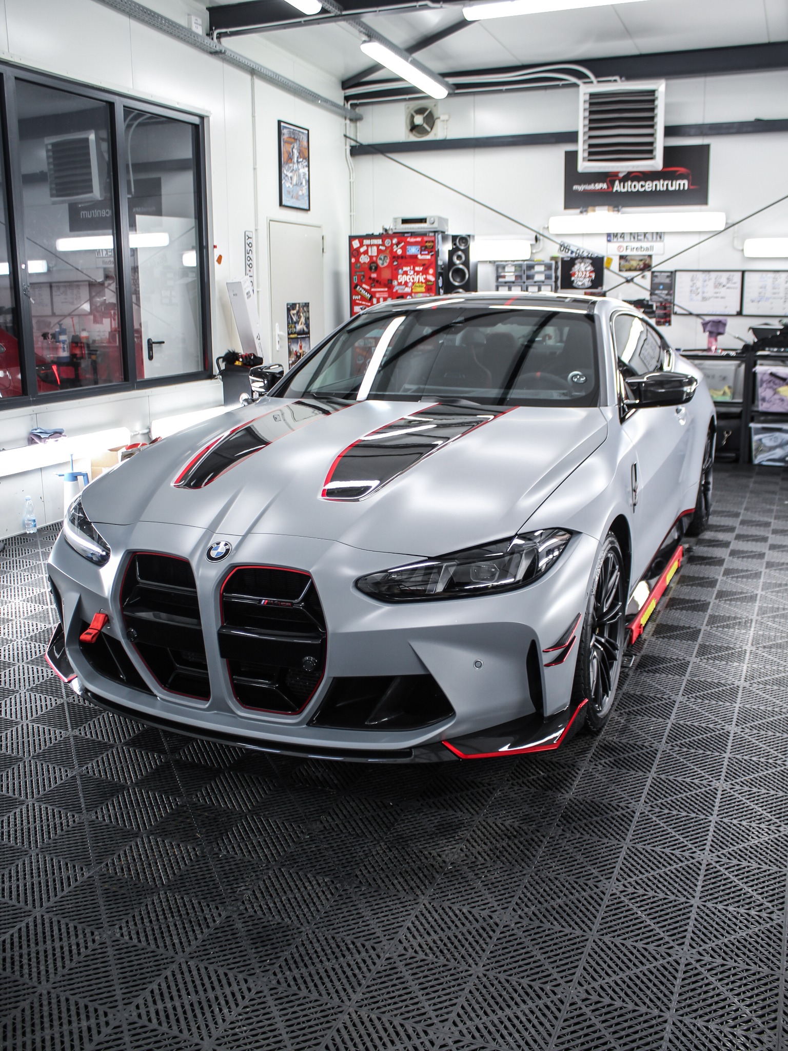 Szary BMW M4 z czarno-czerwonymi akcentami, po zabiegach detailingu, stoi w czystym garażu z oknem i plakatem. Widoczne logo 'myjnia&SPA Autocentrum'.