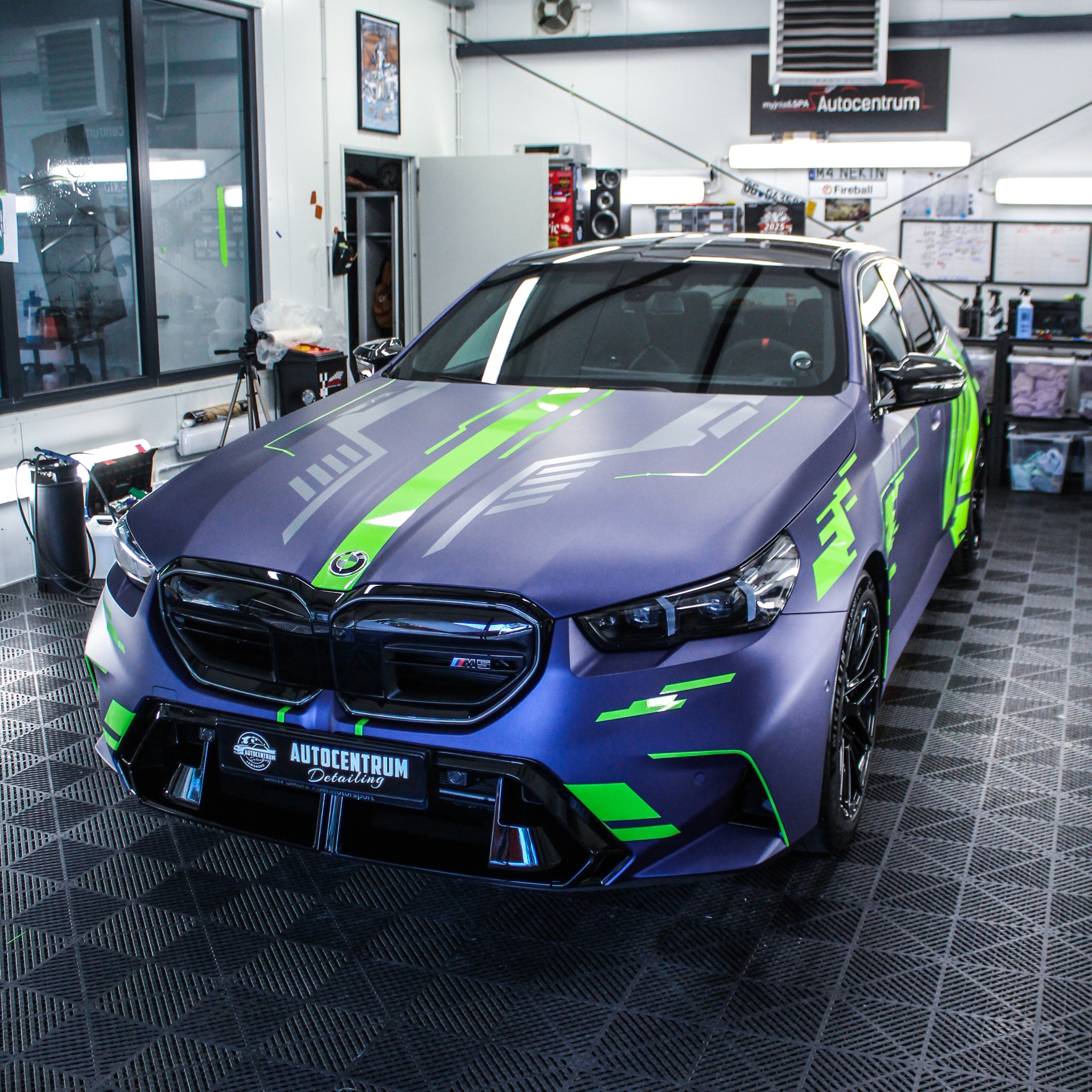 BMW M5 w matowym granacie z neonowymi, zielonymi akcentami graficznymi, zaparkowany w profesjonalnym studiu detailingu. Widok z przodu na oklejoną maskę i zderzak.