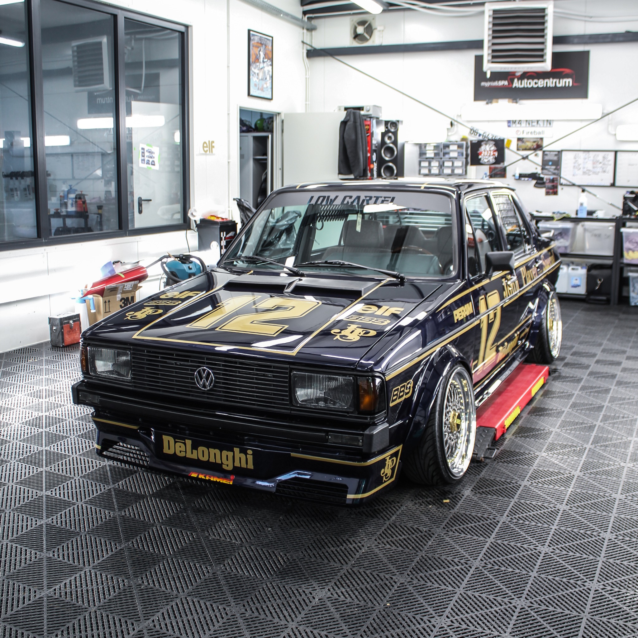 Oklejony, czarny Volkswagen Golf II z numerem 12 w złotym kolorze, stoi w warsztacie. Widoczne liczne logotypy sponsorów i naklejki na karoserii. Efektowny tuning.