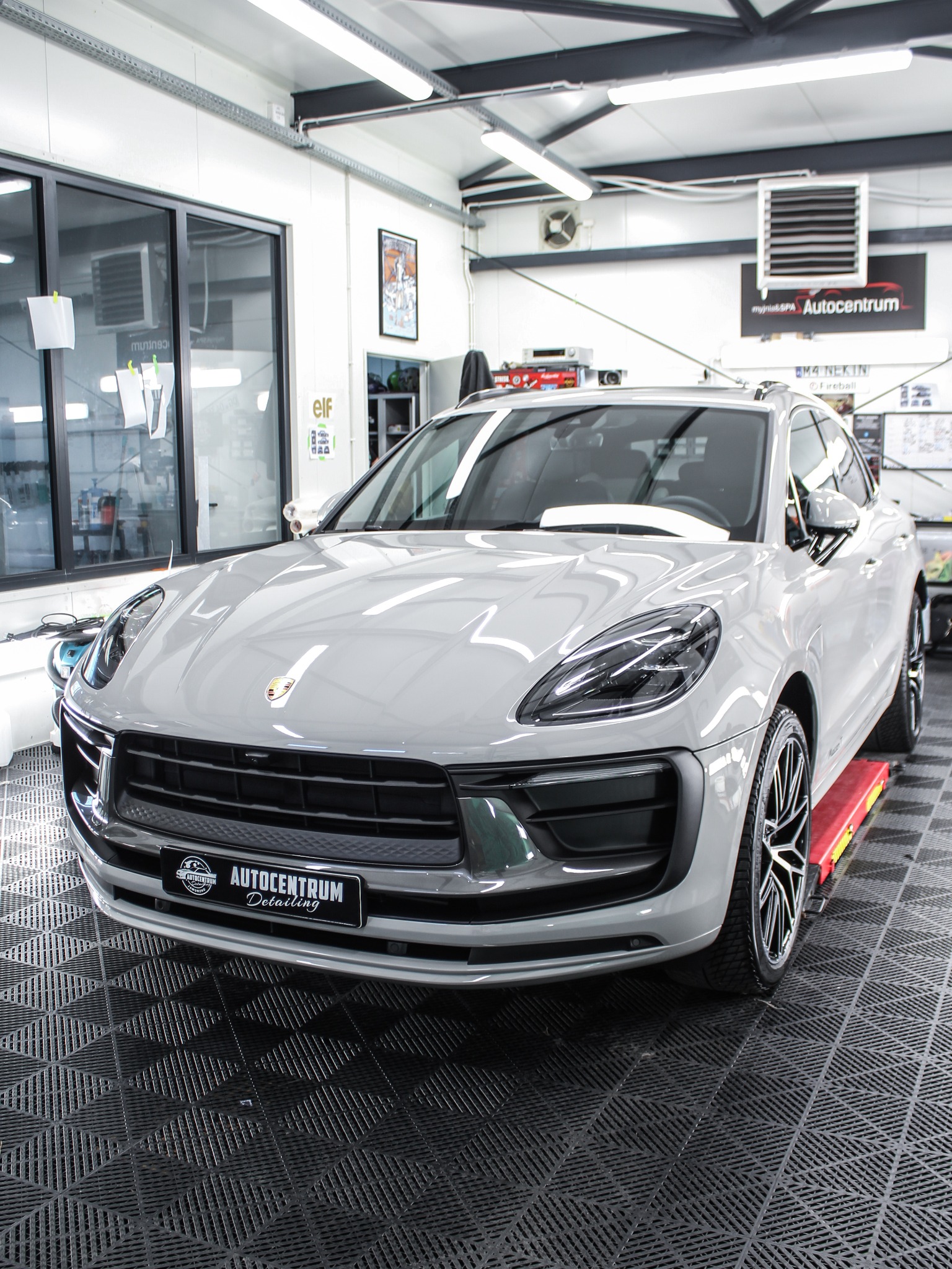 Szary Porsche Macan na podnośniku w warsztacie detailingowym Autocentrum. Widoczne logo firmy na tablicy rejestracyjnej i błyszcząca powłoka lakiernicza.