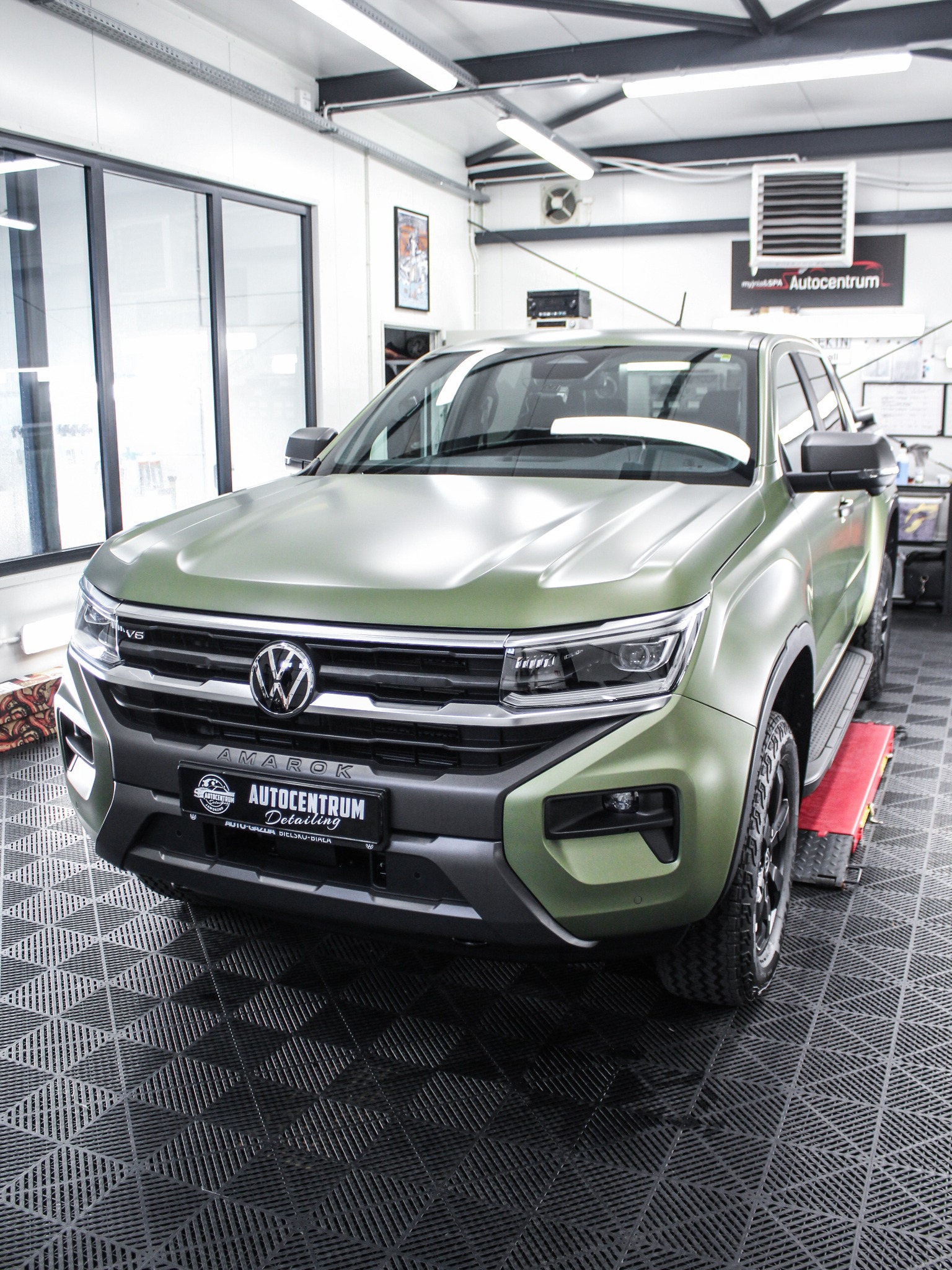 Zielony Volkswagen Amarok po zmianie koloru folią w warsztacie z kratką podłogową i czerwonym podnośnikiem. Widoczny logotyp firmy Autocentrum.