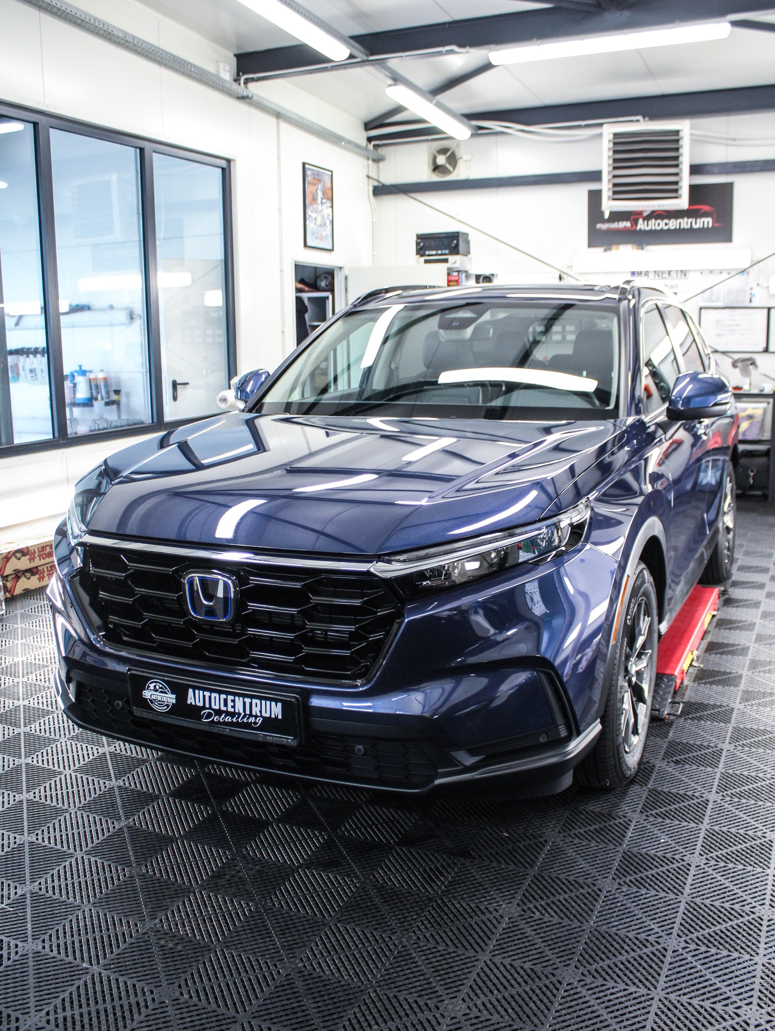 Błyszczący, granatowy samochód Honda na podnośniku w warsztacie detailingowym. Widoczne logo Autocentrum Detailing na tablicy rejestracyjnej i w tle.