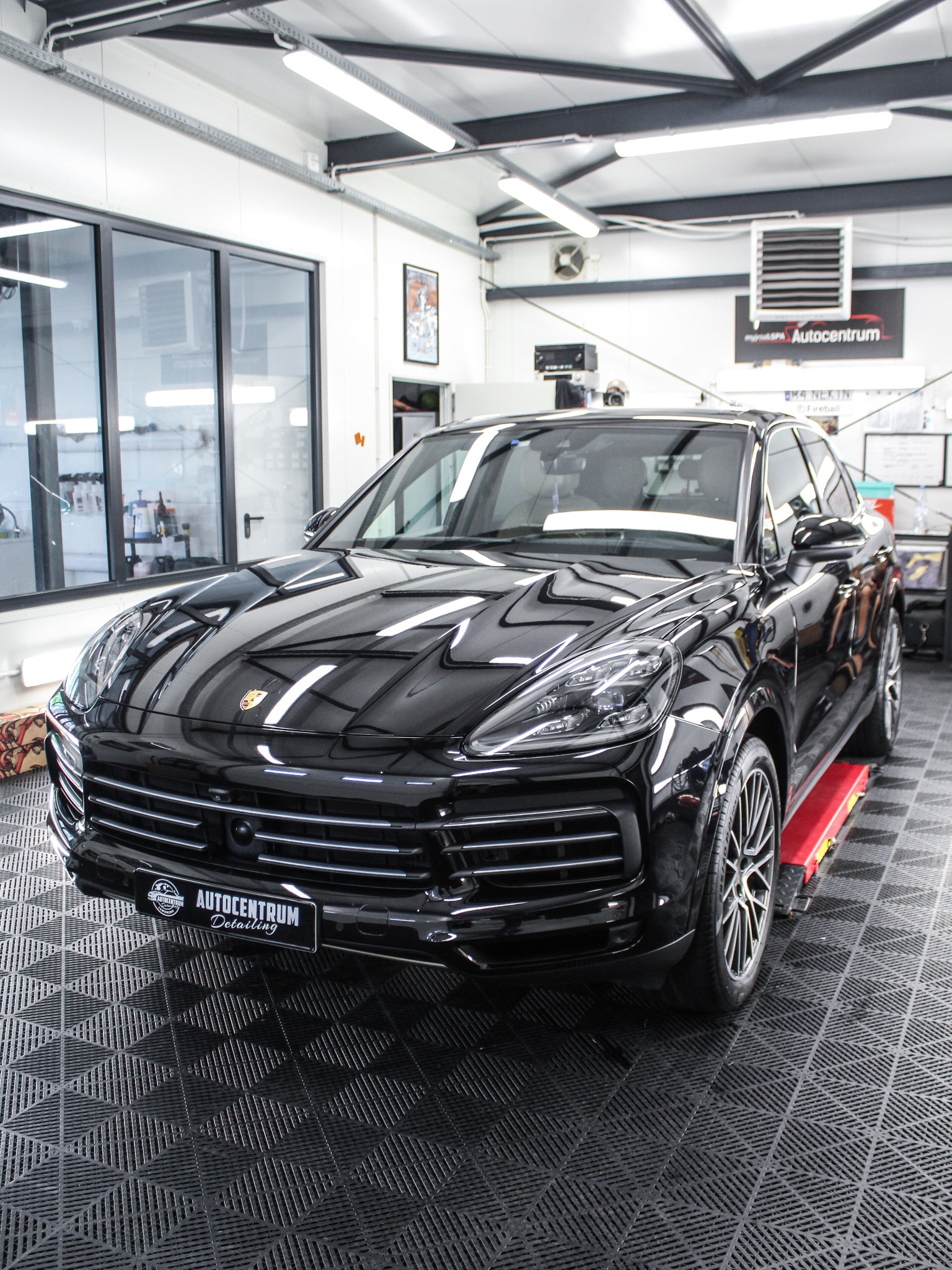 Błyszczący, czarny Porsche Cayenne na podnośniku w dobrze oświetlonym warsztacie detailingowym z logo Autocentrum Detailing na tablicy rejestracyjnej.