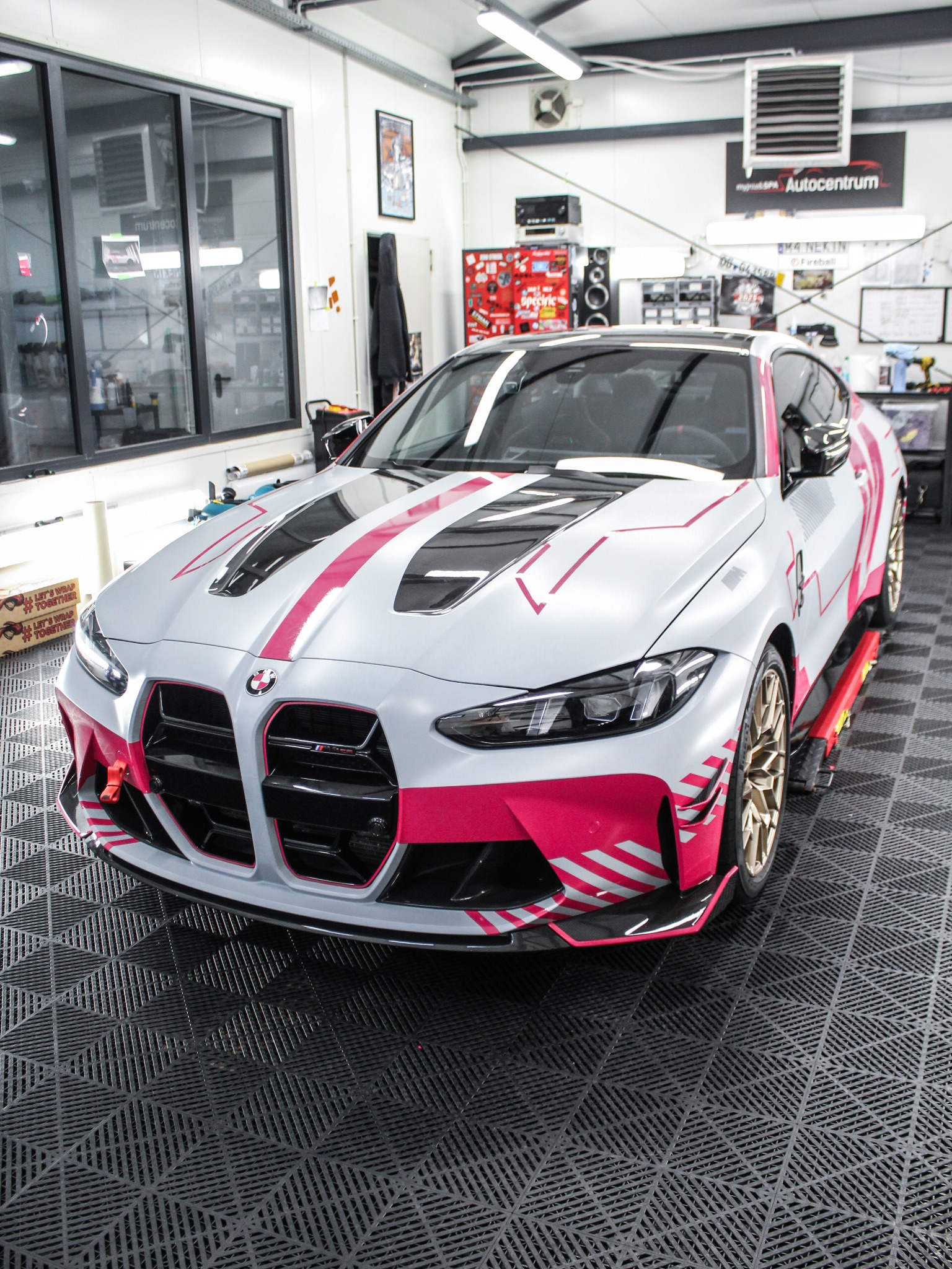 Sportowy BMW M4 w srebrnej folii z różowymi akcentami, zaparkowany w warsztacie na podnośniku. Widoczne detale oklejenia i oświetlenie warsztatowe.