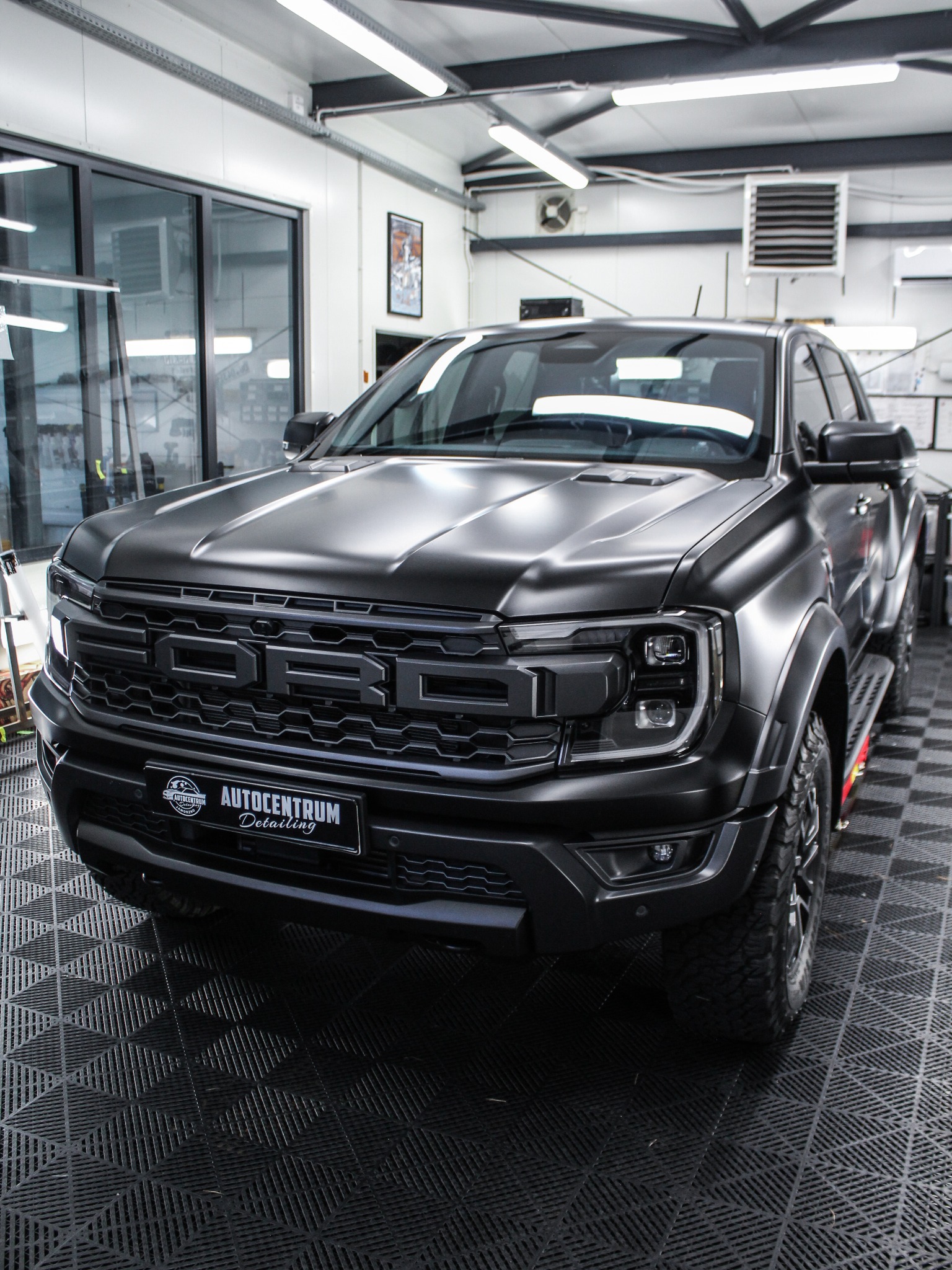 Czarny Ford Raptor po detailingu, z matowym wykończeniem, logo Autocentrum Detailing na tablicy rejestracyjnej, na podłodze z czarnych płytek w garażu.
