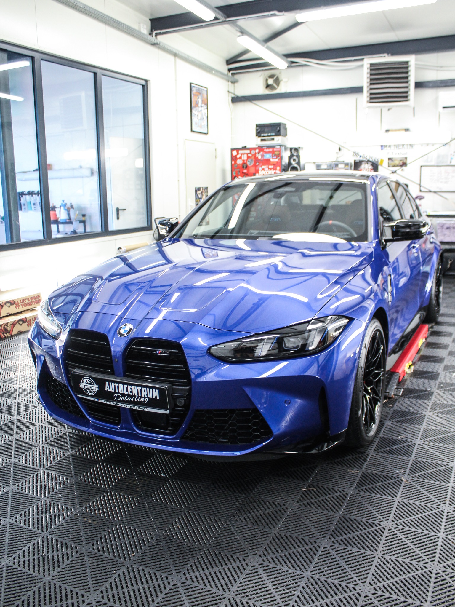 Błyszczący, niebieski BMW M4 na podnośniku w jasnym garażu detailingowym, z widocznym logo Autocentrum Detailing na przednim zderzaku i czarnymi felgami.