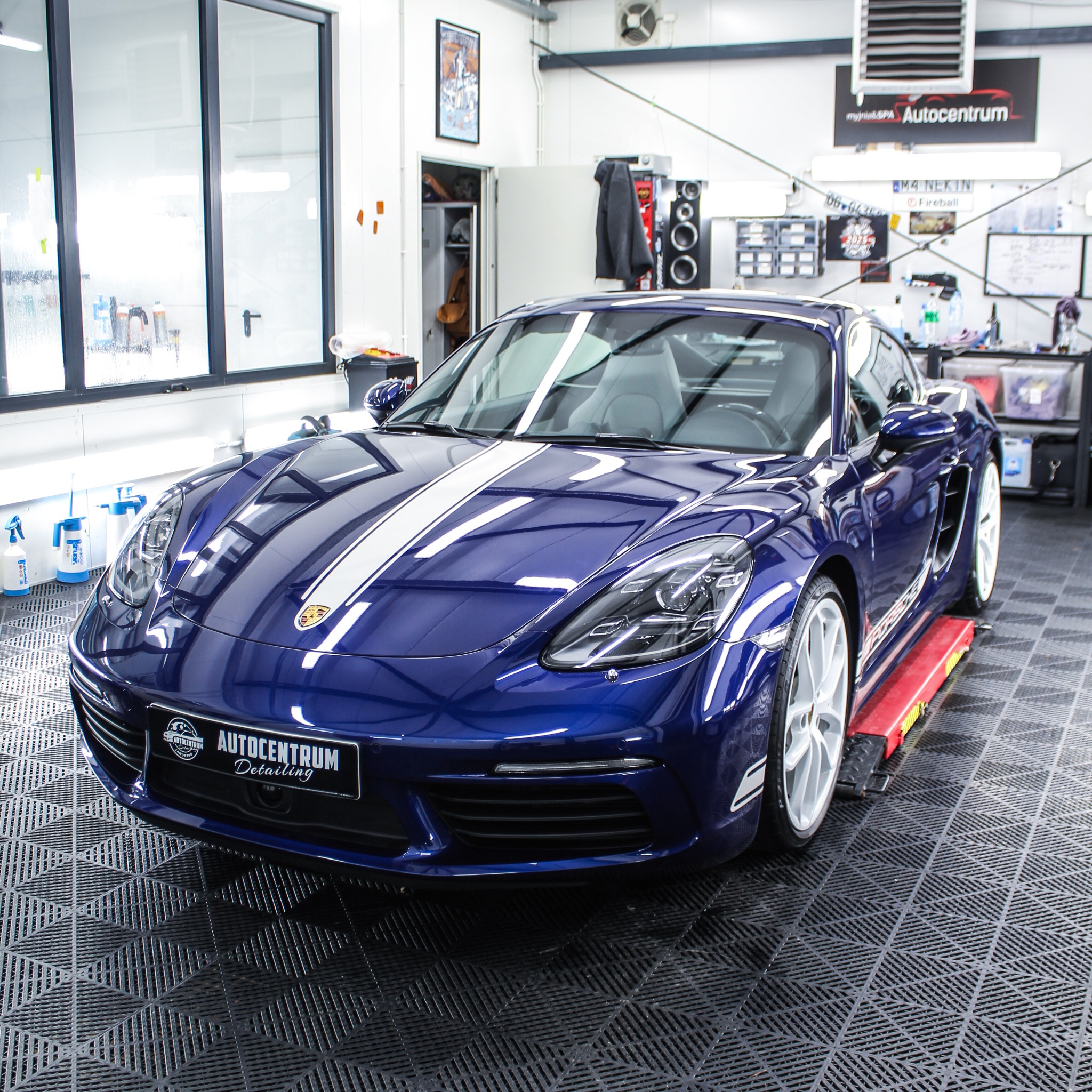 Błękitny Porsche 718 Cayman z białymi pasami na podnośniku w studiu detailingowym, logo Autocentrum Detailing na masce, błyszcząca powłoka lakiernicza.