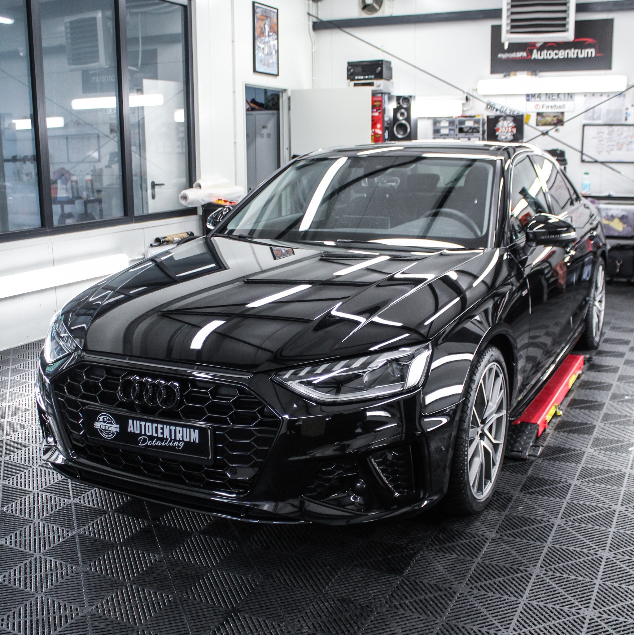 Błyszczący, czarny Audi A4 po detailingu, stoi na czerwonej platformie w czystym, profesjonalnym studiu z oknami i logo Autocentrum Detailing na grillu.
