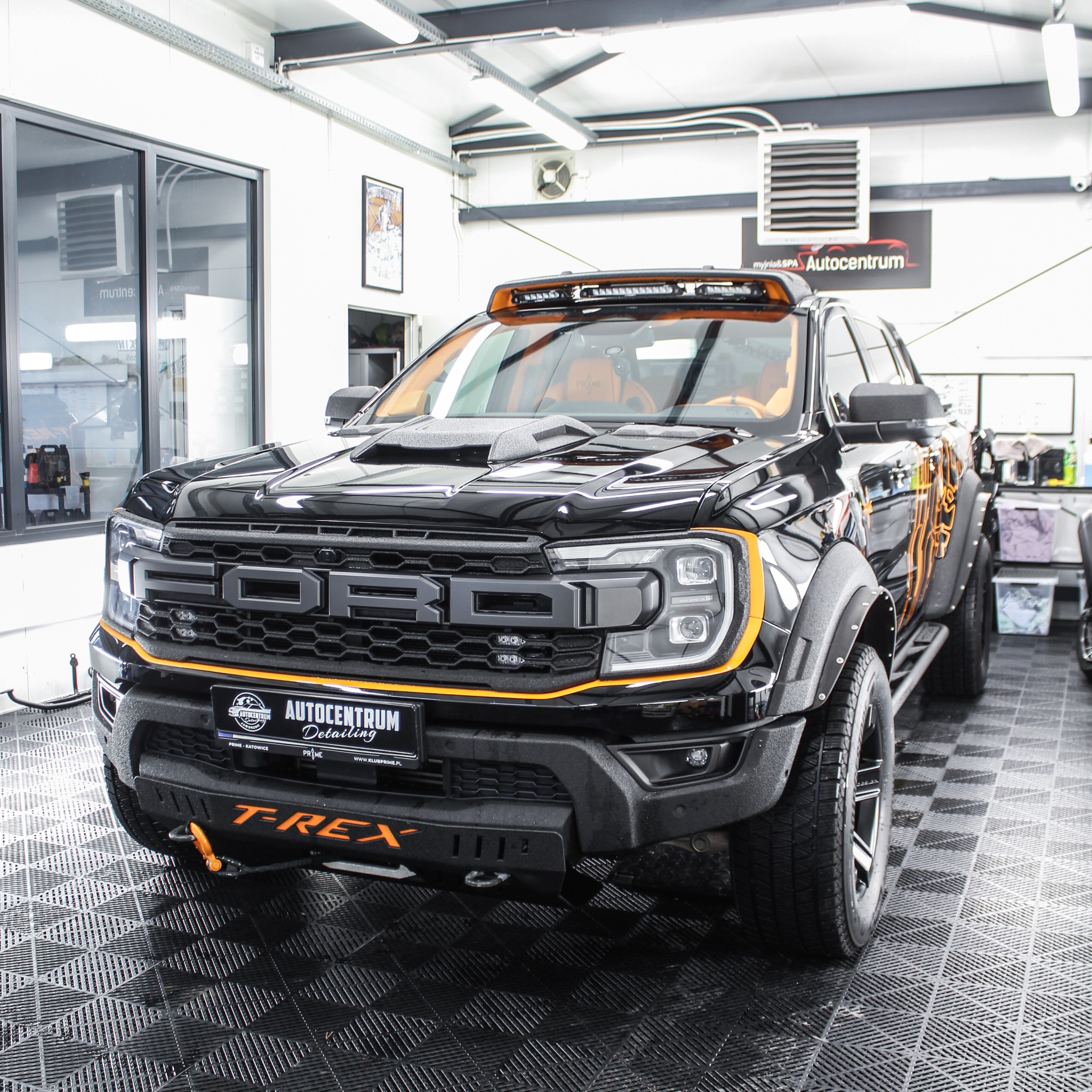 Czarny Ford Ranger Raptor po detailingu z pomarańczowymi akcentami, widok z przodu w jasnym garażu, logo Autocentrum Detailing na tablicy rejestracyjnej.