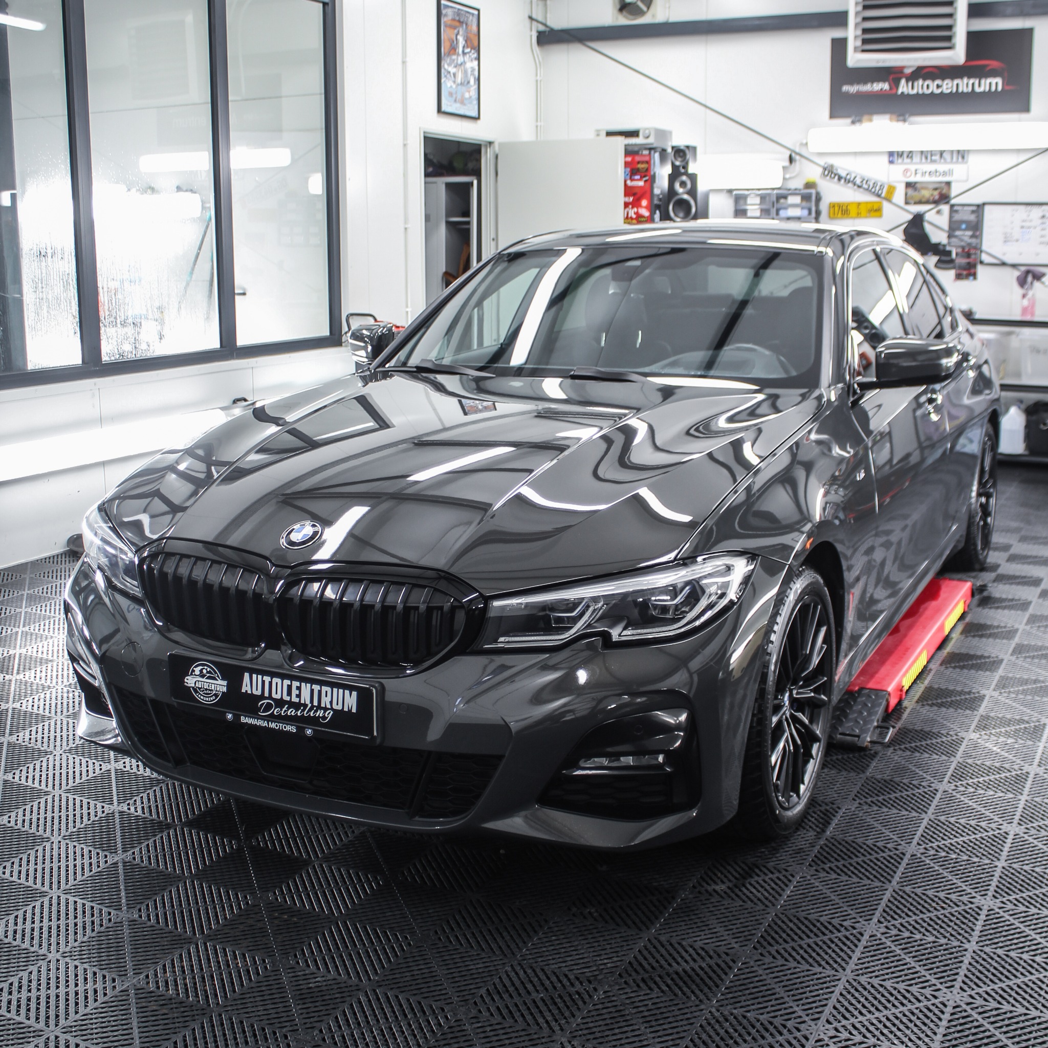 Błyszczący, szary samochód BMW po detailingu, ustawiony na podnośniku w sterylnym garażu. Widoczne logo Autocentrum Detailing i odbicia w lakierze.