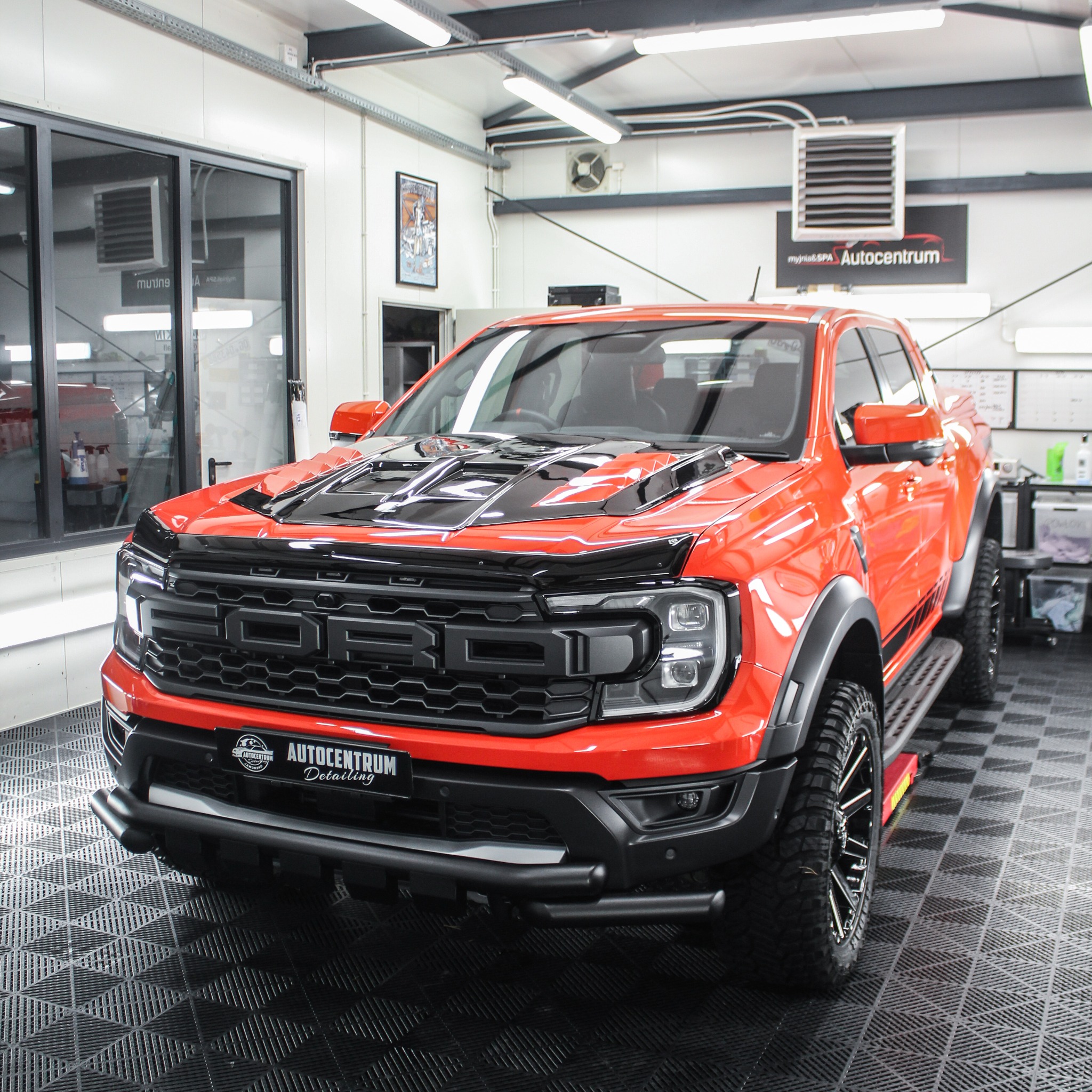 Pomarańczowy pickup Ford Ranger Raptor po detailingu, z czarnymi akcentami na masce i grillu, zaparkowany w czystym, dobrze oświetlonym garażu detailingowym.