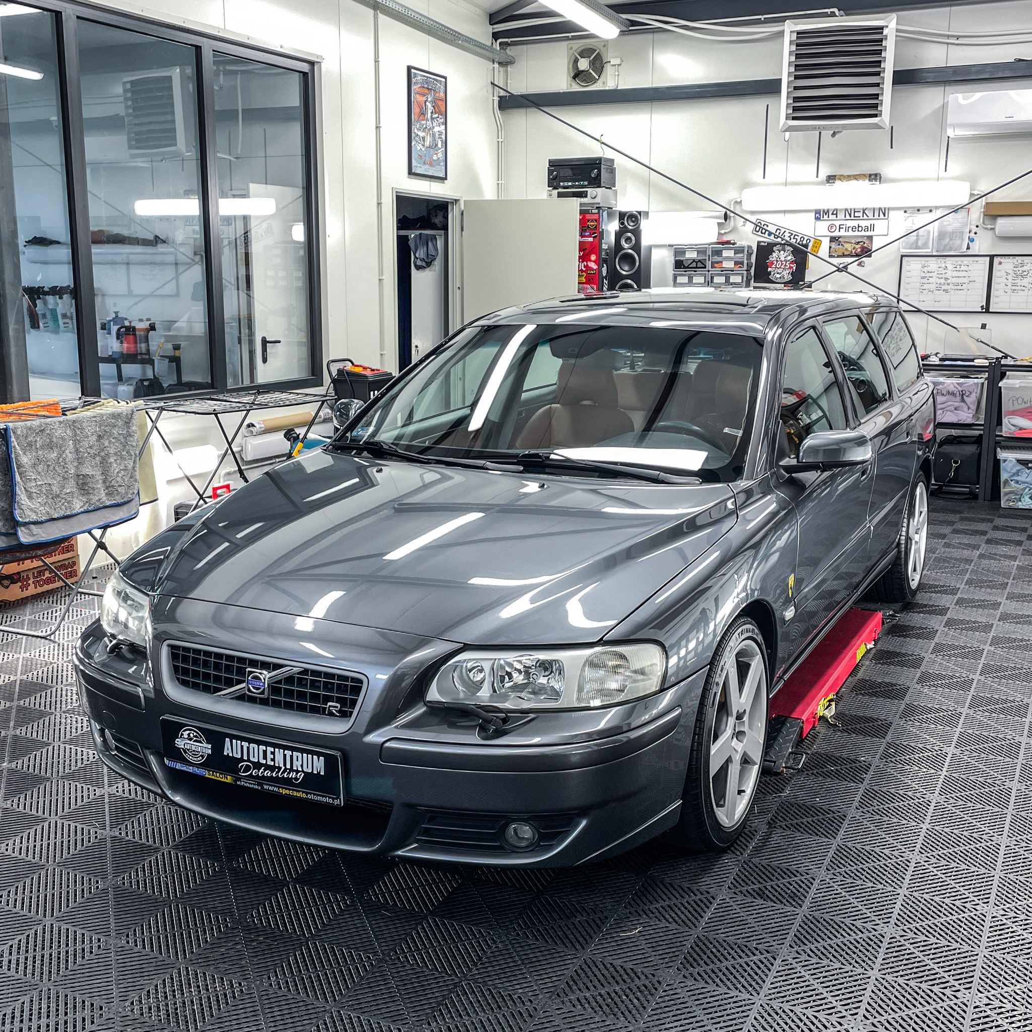 Szary samochód Volvo V70 na podnośniku w dobrze oświetlonym garażu detailingowym z wyposażeniem i materiałami do pielęgnacji aut.