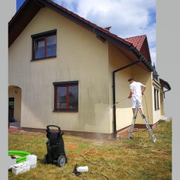 Rensar Polska - Pracownik na drabinie czyści elewację domu za pomocą myjki ciśnieniowej. Widoczne zabrudzenia na ścianie przed czyszczeniem i czysty fragment po umyciu. Na trawie leży sprzęt.