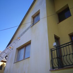 Rensar Polska - Żółta elewacja domu po umyciu, widoczne zacieki i drabina oparta o ścianę, błękitne niebo w tle. Częściowo umyta ściana budynku z balkonem.