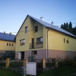 Żółty dom dwukondygnacyjny z balkonem i szarą dachówką, otoczony czarnym, kutym ogrodzeniem. Część elewacji pokryta dekoracyjną okładziną. W tle zieleń i błękitne niebo.