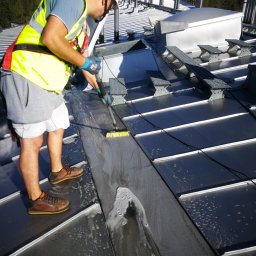 Rensar Polska - Pracownik w kasku i kamizelce czyści szczotką metalowy dach przemysłowy, widoczne mydliny i częściowo umyta powierzchnia. W tle drzewa i elementy konstrukcji.