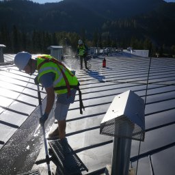 Rensar Polska - Pracownik w kasku i kamizelce myje metalowy dach za pomocą szczotki na kiju. W tle góry i drugi pracownik. Widoczne elementy wentylacji dachowej.