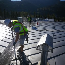 Rensar Polska - Ekipa w kaskach i kamizelkach czyści metalowy dach z użyciem szczotek i wody. W tle góry i błękitne niebo.