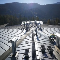 Rensar Polska - Praca na dachu z blachy na tle gór. Dwóch pracowników w kamizelkach odblaskowych przy metalowych elementach wentylacyjnych. Widoczne drabinki dachowe.