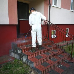 Rensar Polska - Pracownik w kombinezonie czyści ciśnieniowo schody wejściowe z czerwono-szarej kostki, z widocznymi zabrudzeniami i strumieniem wody. Budynek z elewacją w odcieniach beżu i czerwieni.