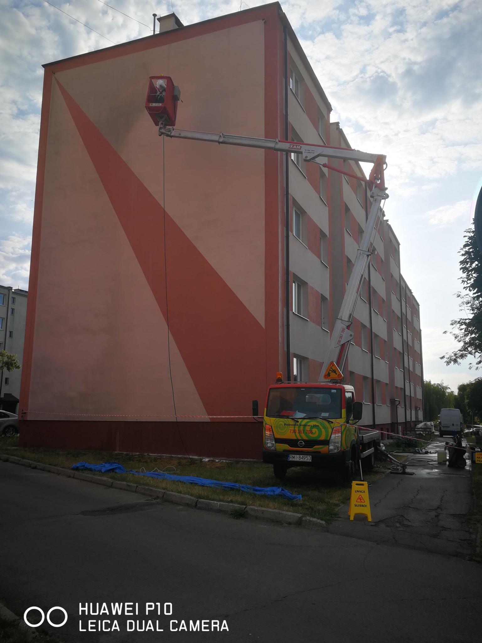 Pracownik w koszu podnośnika czyści elewację budynku z geometrycznym wzorem. Żółty samochód z podnośnikiem koszowym zaparkowany obok.