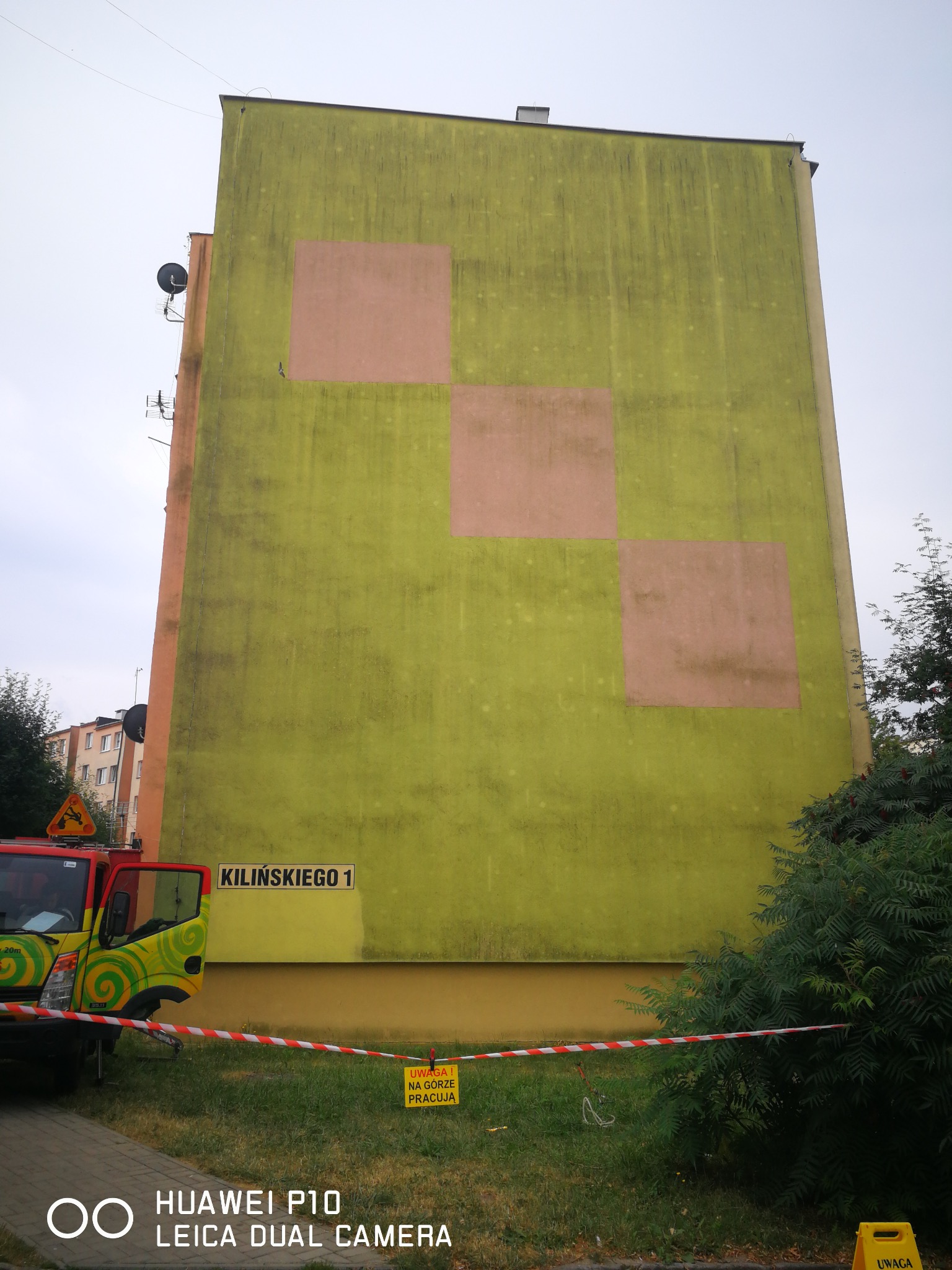 Blok z zieloną elewacją, na której widoczne są kwadraty w innym odcieniu. Tabliczka z adresem 'Kilińskiego 1'. Taśma ostrzegawcza i znak 'Uwaga! Na górze pracują'.