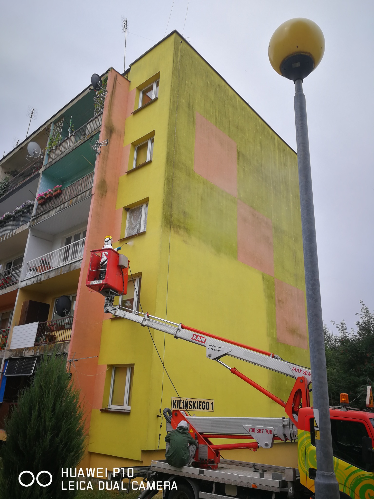 Pracownik w podnośniku koszowym myje elewację bloku. Widoczne zielone zabrudzenia. Żółto-brzoskwiniowa fasada. Ulica Kilińskiego 1. Numer telefonu na pojeździe.