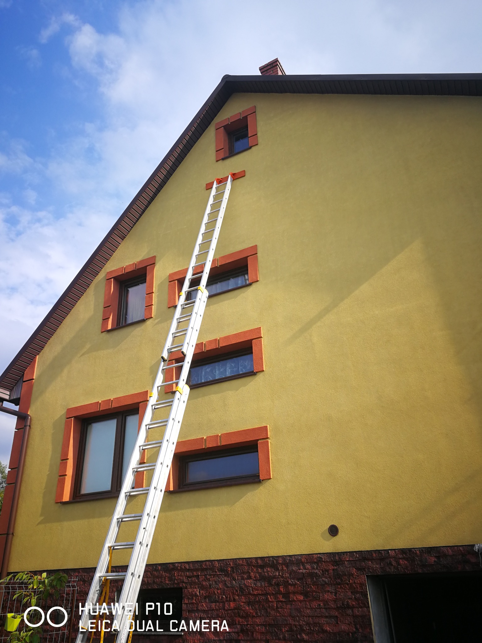 Żółta elewacja domu z ceglaną podmurówką i oknami w pomarańczowych ramach. Do ściany przystawiona długa, aluminiowa drabina. Błękitne niebo z białymi chmurami w tle.