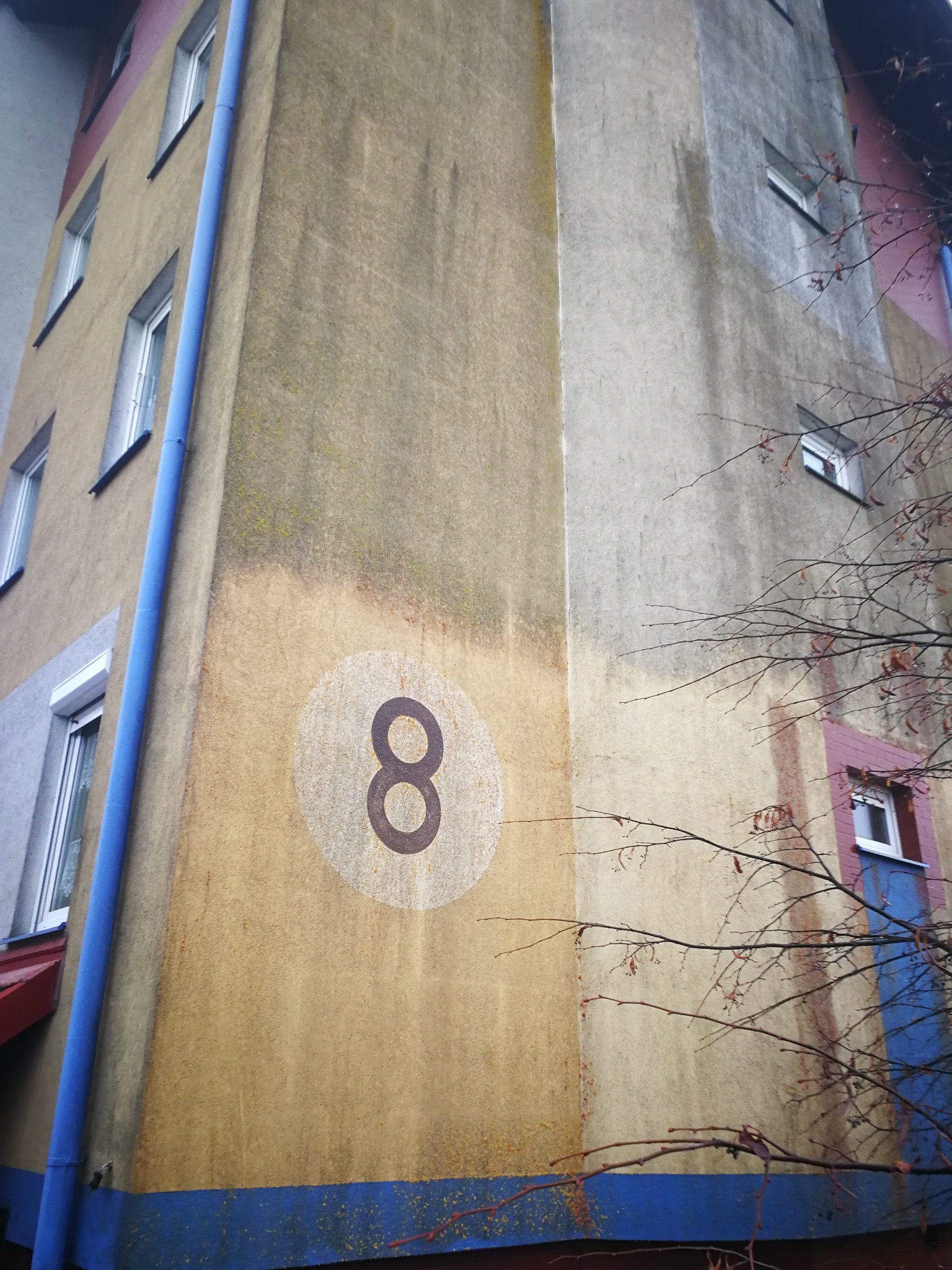 Wielopiętrowy budynek z numerem osiem na elewacji, widoczne okna i rynna. Elewacja częściowo zabrudzona, z widocznymi śladami zacieków. Ujęcie z dołu, z fragmentem drzewa.