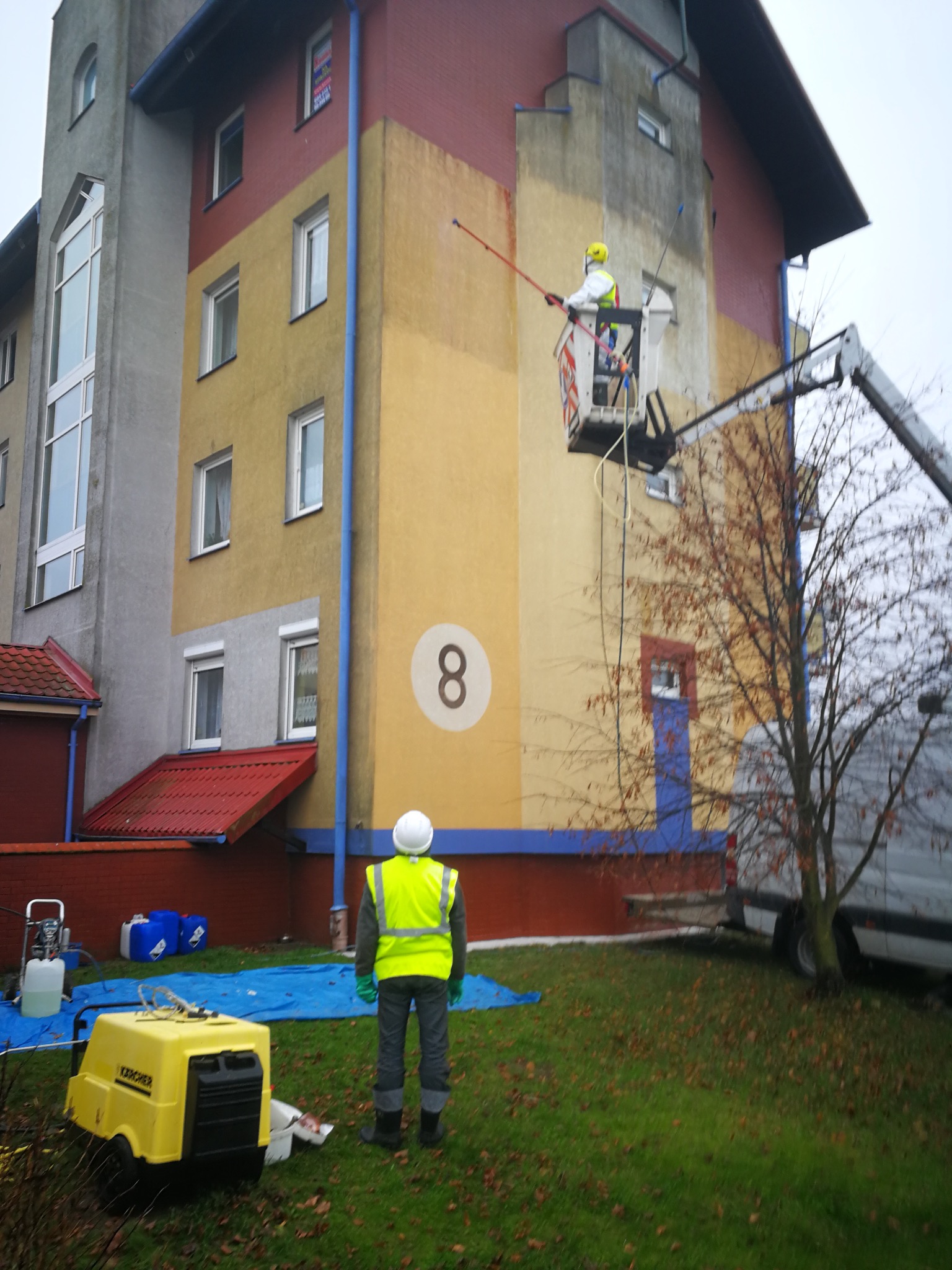 Pracownik na podnośniku czyści elewację budynku z numerem '8'. Na trawniku stoi żółta myjka ciśnieniowa Karcher oraz drugi pracownik w kamizelce.