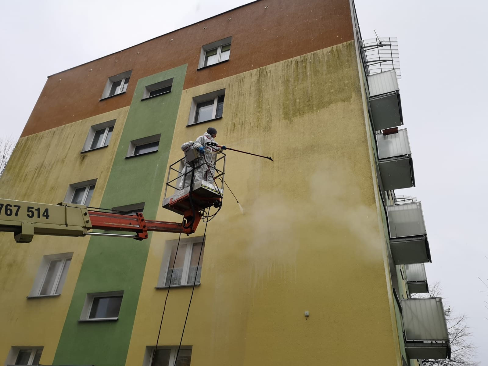 Pracownik w kombinezonie czyści elewację budynku wielorodzinnego z podnośnika koszowego za pomocą myjki ciśnieniowej. Widoczne zabrudzenia na fasadzie.