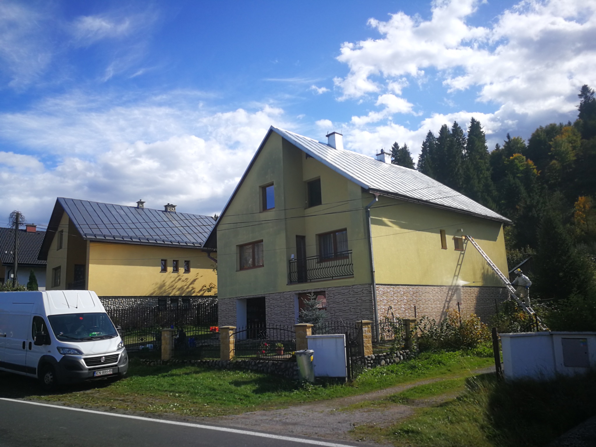 Dwupiętrowy dom z żółtą elewacją, czyszczony przez pracownika na drabinie. W tle drugi dom i las, a z przodu biały van. Błękitne niebo z chmurami.