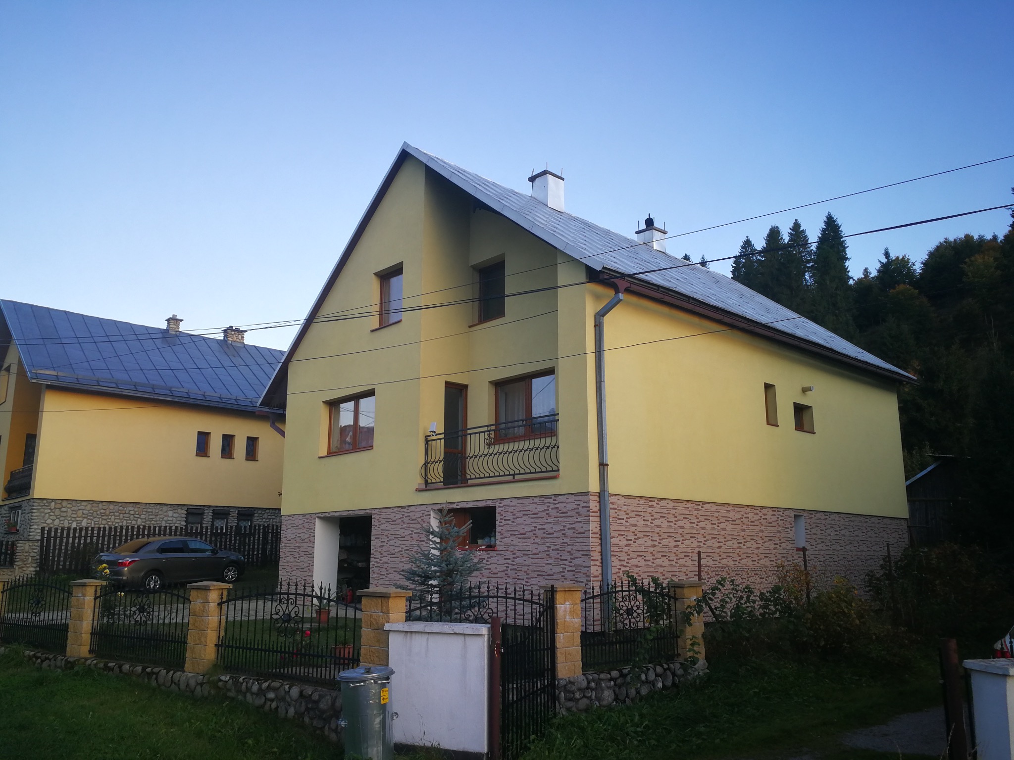Żółty dom dwukondygnacyjny z balkonem i szarą dachówką, otoczony czarnym, kutym ogrodzeniem. Część elewacji pokryta dekoracyjną okładziną. W tle zieleń i błękitne niebo.