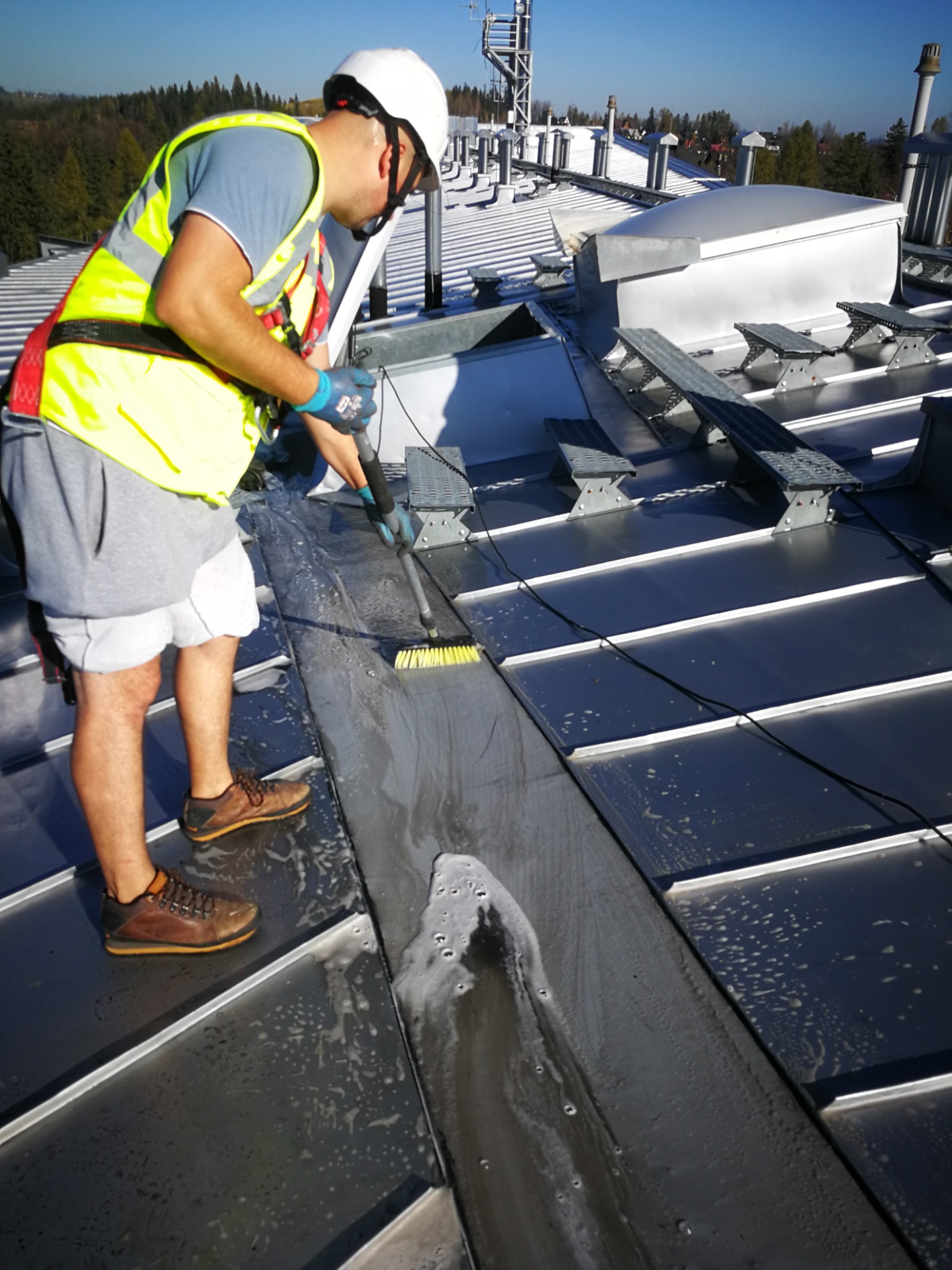 Pracownik w kasku i kamizelce czyści szczotką metalowy dach przemysłowy, widoczne mydliny i częściowo umyta powierzchnia. W tle drzewa i elementy konstrukcji.