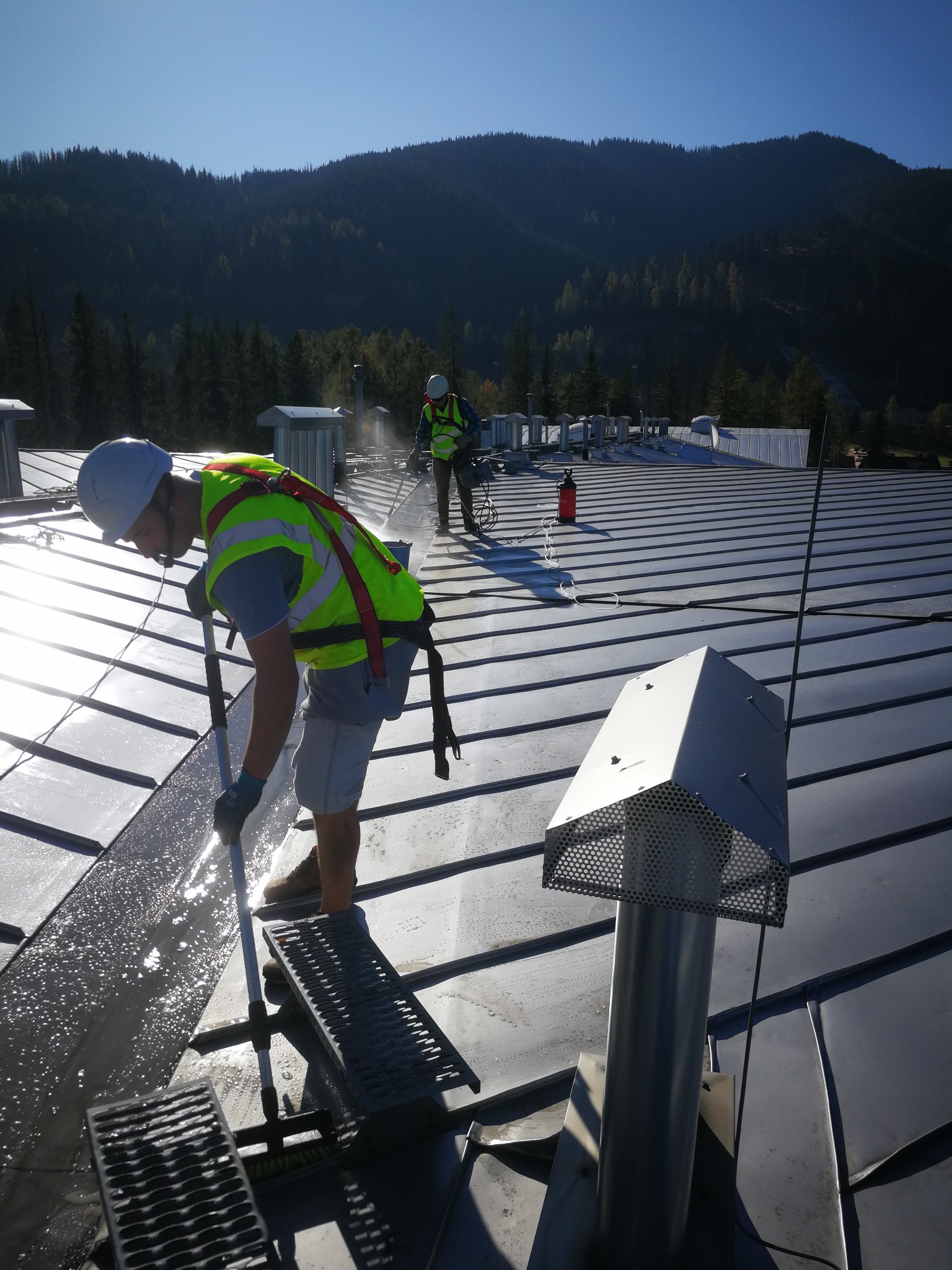 Pracownik w kasku i kamizelce myje metalowy dach za pomocą szczotki na kiju. W tle góry i drugi pracownik. Widoczne elementy wentylacji dachowej.