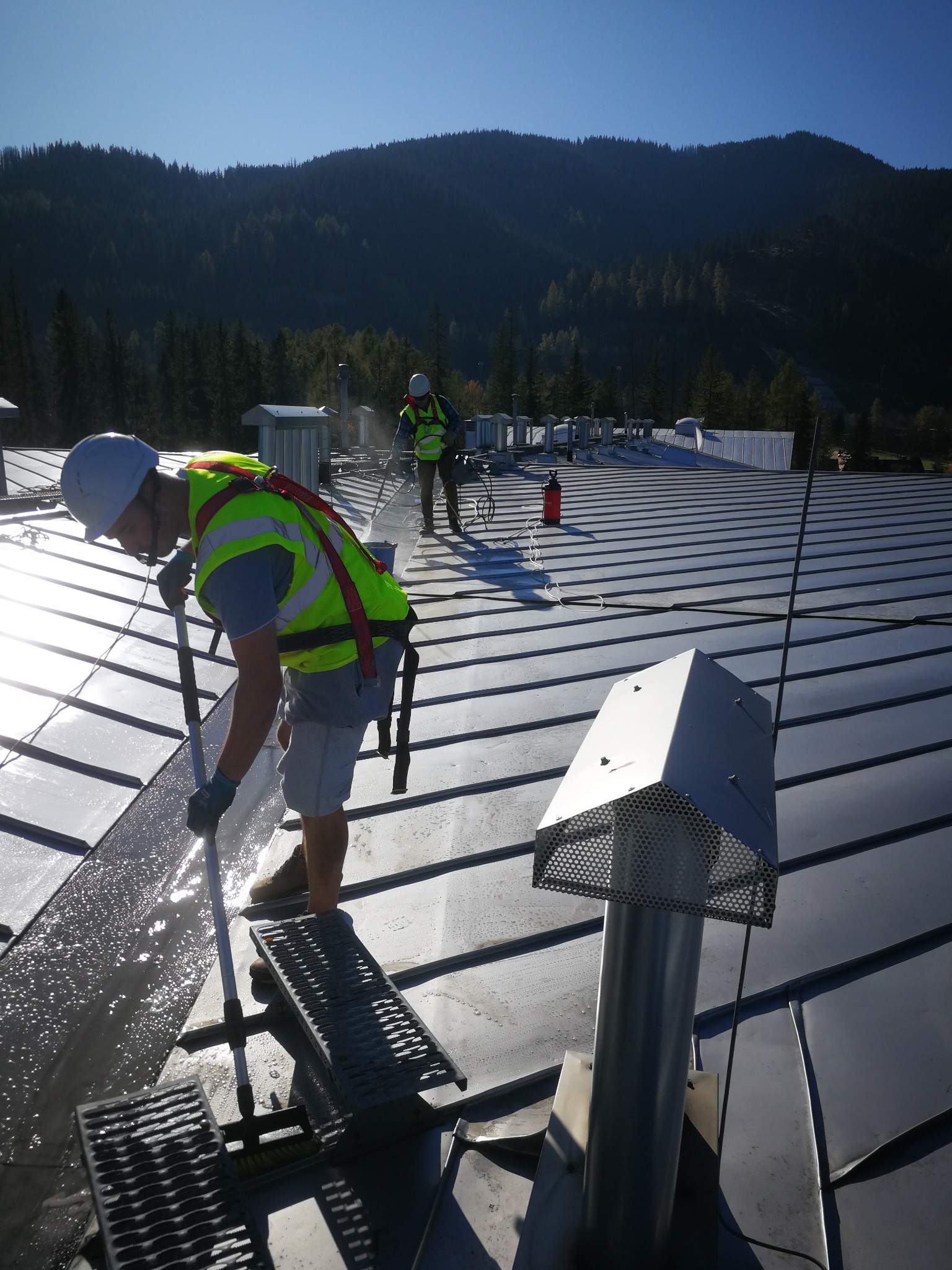 Ekipa w kaskach i kamizelkach czyści metalowy dach z użyciem szczotek i wody. W tle góry i błękitne niebo.
