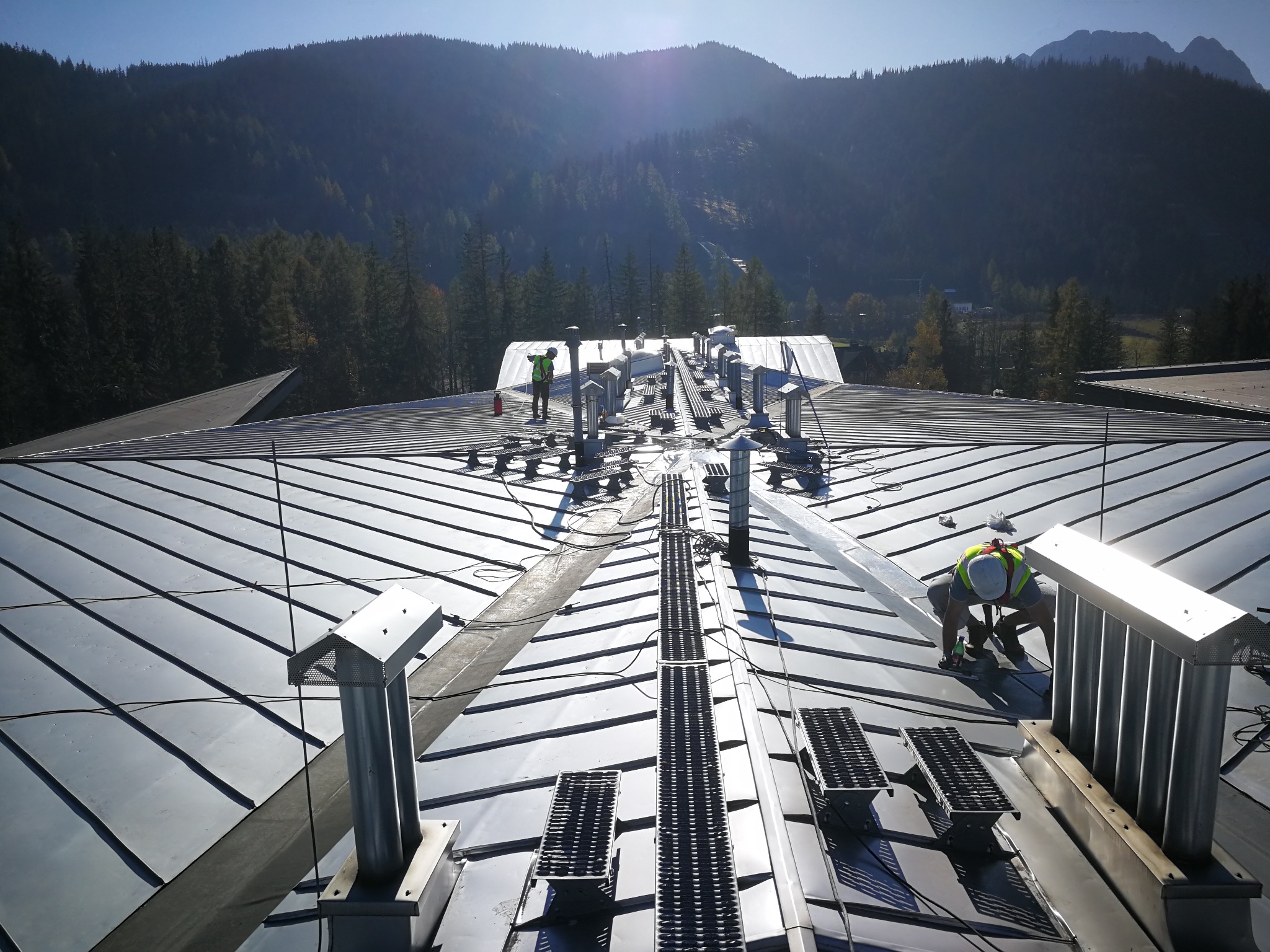 Praca na dachu z blachy na tle gór. Dwóch pracowników w kamizelkach odblaskowych przy metalowych elementach wentylacyjnych. Widoczne drabinki dachowe.