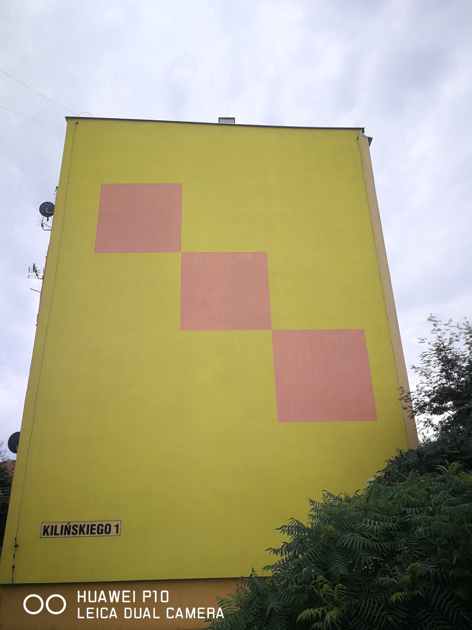 Żółta elewacja bloku z trzema różowymi kwadratami, widok z dołu, tabliczka z adresem 'Kilińskiego 1', satelity na ścianie, fragment zieleni.