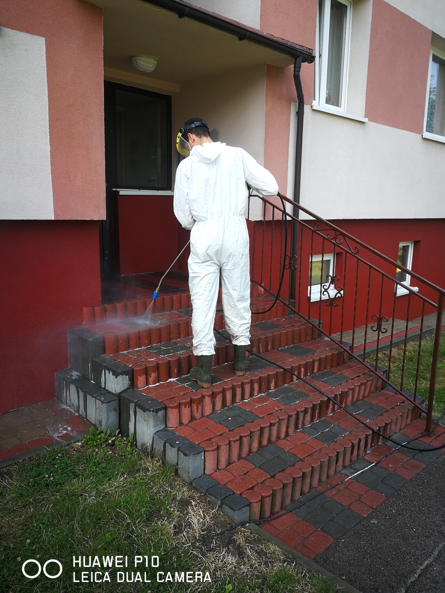Pracownik w kombinezonie czyści ciśnieniowo schody wejściowe z czerwono-szarej kostki, z widocznymi zabrudzeniami i strumieniem wody. Budynek z elewacją w odcieniach beżu i czerwieni.
