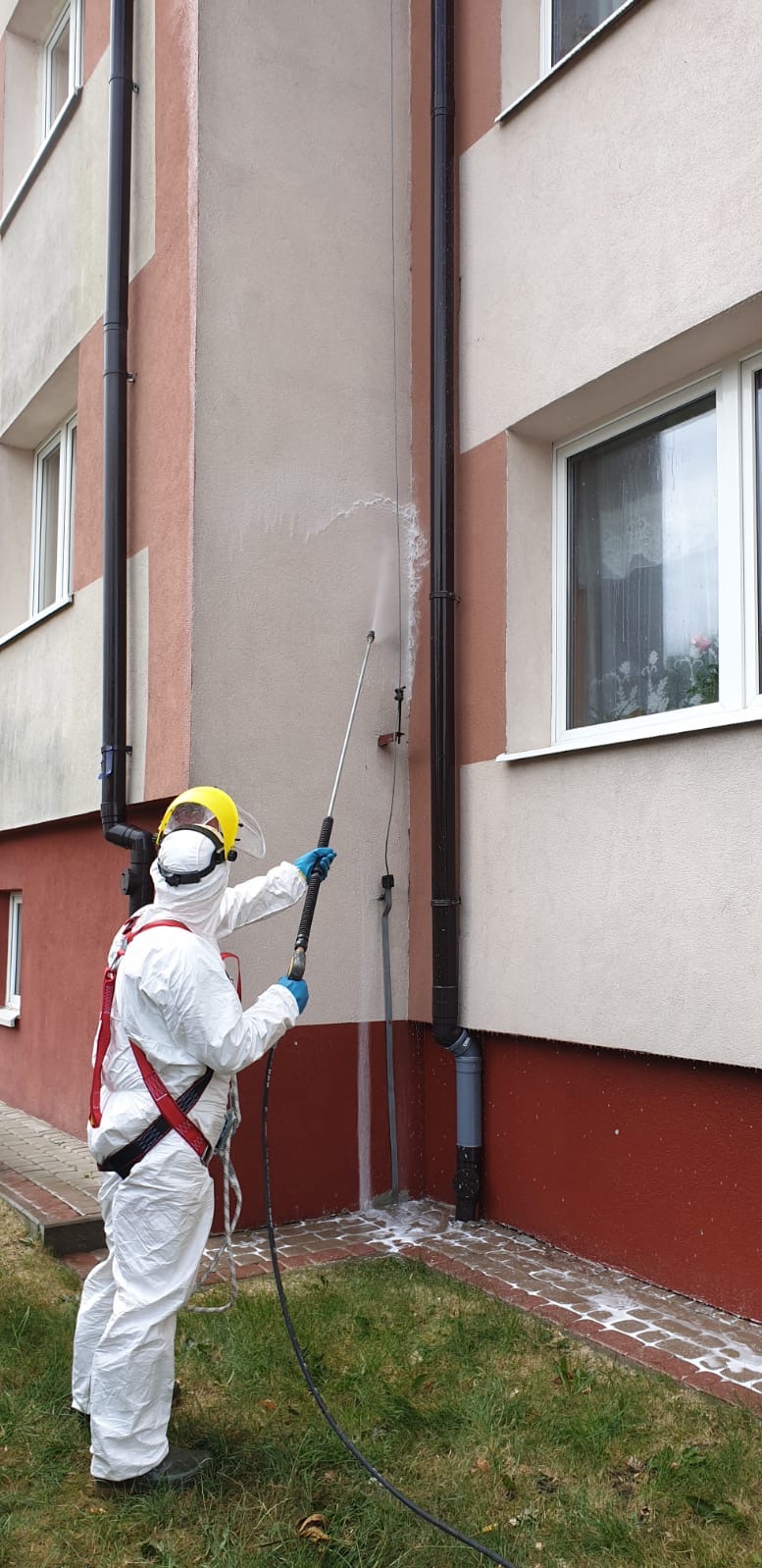 Pracownik w kombinezonie ochronnym czyści elewację budynku z użyciem myjki ciśnieniowej. Widoczne zacieki i piana po czyszczeniu.