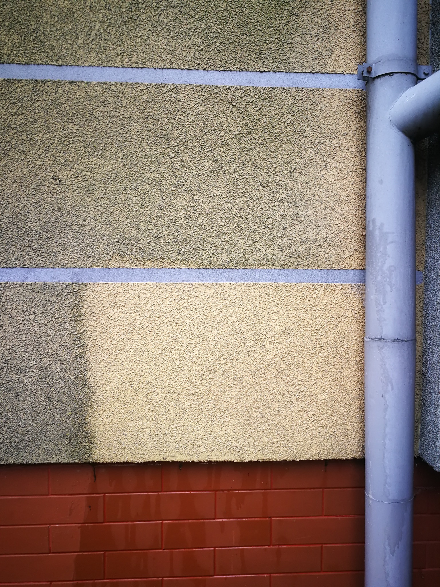 Fragment elewacji z widocznym zabrudzeniem na górnej części i rurą spustową. Dolna część elewacji wykończona czerwoną cegłą.
