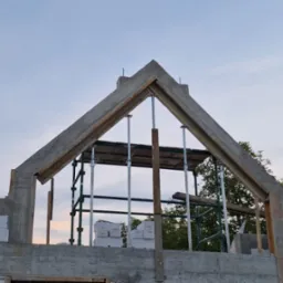 Wznoszenie betonowej konstrukcji domu jednorodzinnego. Widoczne szalunki i podpory stropu. Ściany z betonu komórkowego. Budowa w toku przy pogodnym niebie.